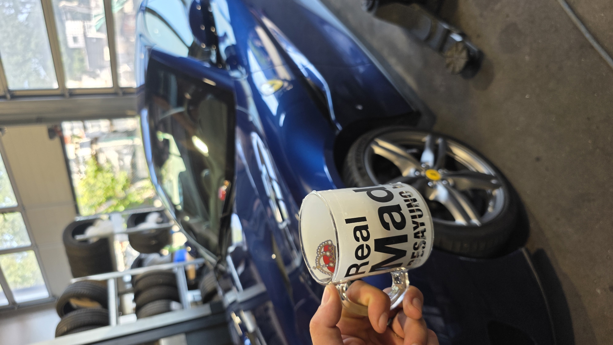 A person's hand holds a glass mug with text that reads "Real Mad Desayuno" and a crown graphic. The mug is held in front of a blue sports car. The car is parked in what appears to be a garage or repair shop, with shelves of tires visible in the background. A car lift is also present in the scene. The lighting suggests an indoor setting, possibly with natural light coming from a large garage door.
