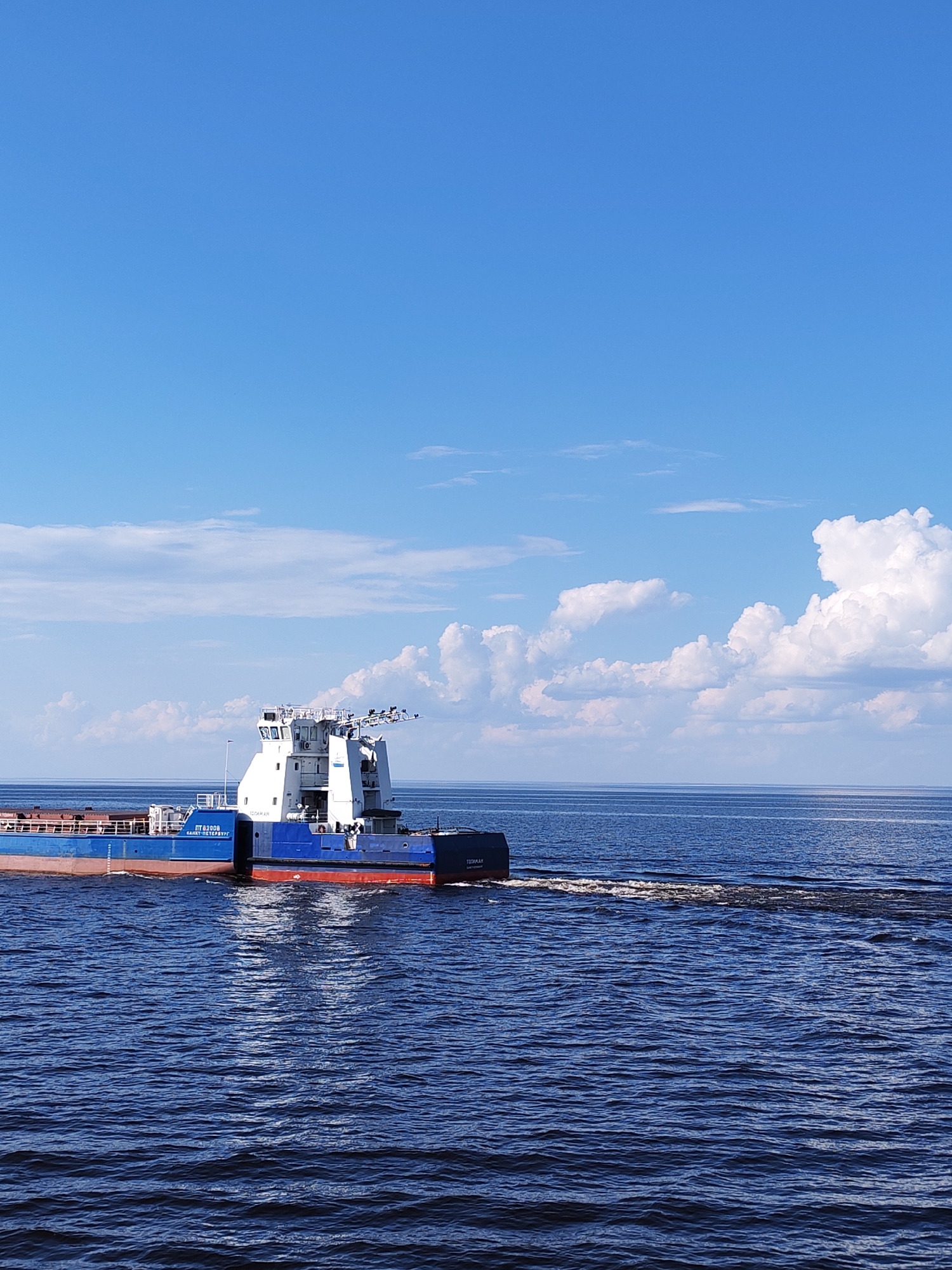 The image shows a blue and red barge traveling on the dark blue water of what appears to be a large body of water, likely a river or lake, with a clear blue sky above dotted with white clouds. The barge is a flat-bottomed vessel with a white superstructure towards the back. The wake of the barge is visible in the water behind it. The words "Saint-Petersburg" are printed on the barge. The city of Veregonets, Russia is not apparent in this photograph.