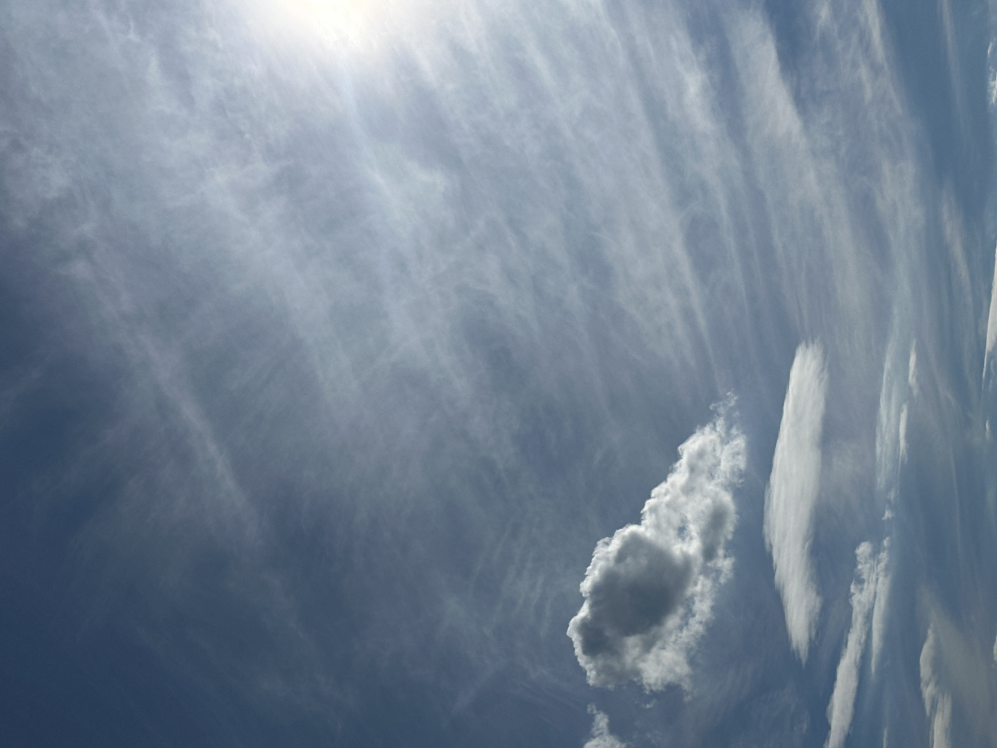 The image captures a dynamic sky filled with wispy, elongated clouds that stretch diagonally across the frame. The sunlight, originating from the upper left corner, is bright and diffused, creating a hazy, luminous effect that bathes the sky in a soft glow. This suggests it is likely daytime, possibly late morning or early afternoon, when the sun is high.
Dominating the lower right portion of the image is a distinct, puffy cumulus cloud, appearing denser and more substantial than the surrounding cirrus formations. To its left, another smaller, darker cumulus cloud is visible. The contrast between the brighter, feathery clouds and the more solid cumulus clouds adds depth and texture to the sky. The overall impression is one of a serene and somewhat dramatic natural display.
The scene is an outdoor setting, specifically a view looking upwards at the sky. There are no discernible people, objects, or text present in the image. The weather appears to be partly cloudy, with a mix of high, wispy clouds and lower, more defined clouds, under bright sunlight. The overall mood is one of openness and natural beauty.