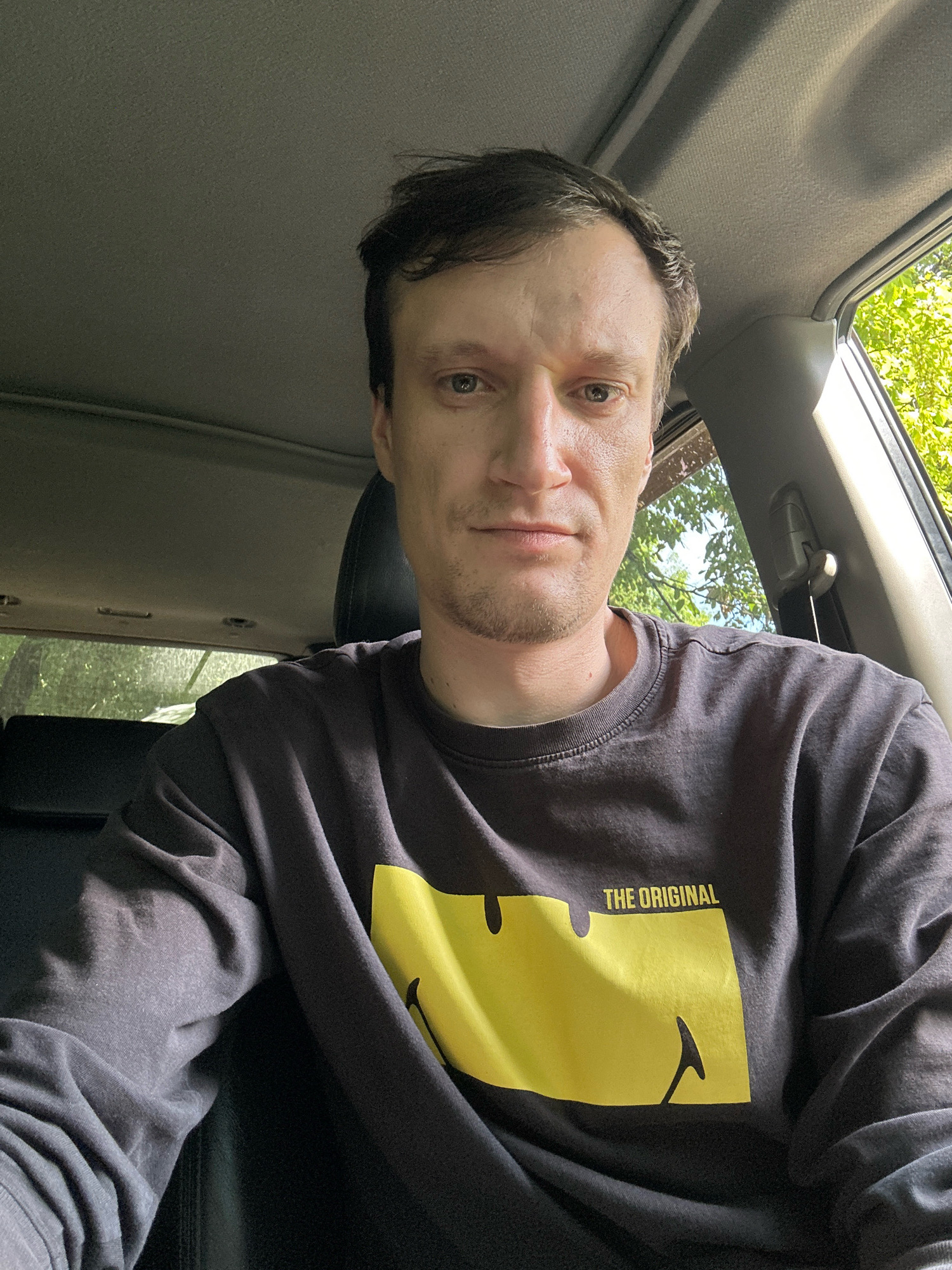 A man is taking a selfie from the driver's seat of a car. He has short brown hair and is wearing a dark long-sleeved shirt with a yellow smiley face graphic and the words "THE ORIGINAL" printed on it. The car interior is visible, including the headliner, a sunroof, and part of the passenger seat. Through the windows, green trees and foliage are visible, suggesting an outdoor, possibly rural or park-like setting. The image is taken during the daytime, with sunlight illuminating the scene and casting shadows. The overall impression is a candid, casual shot.