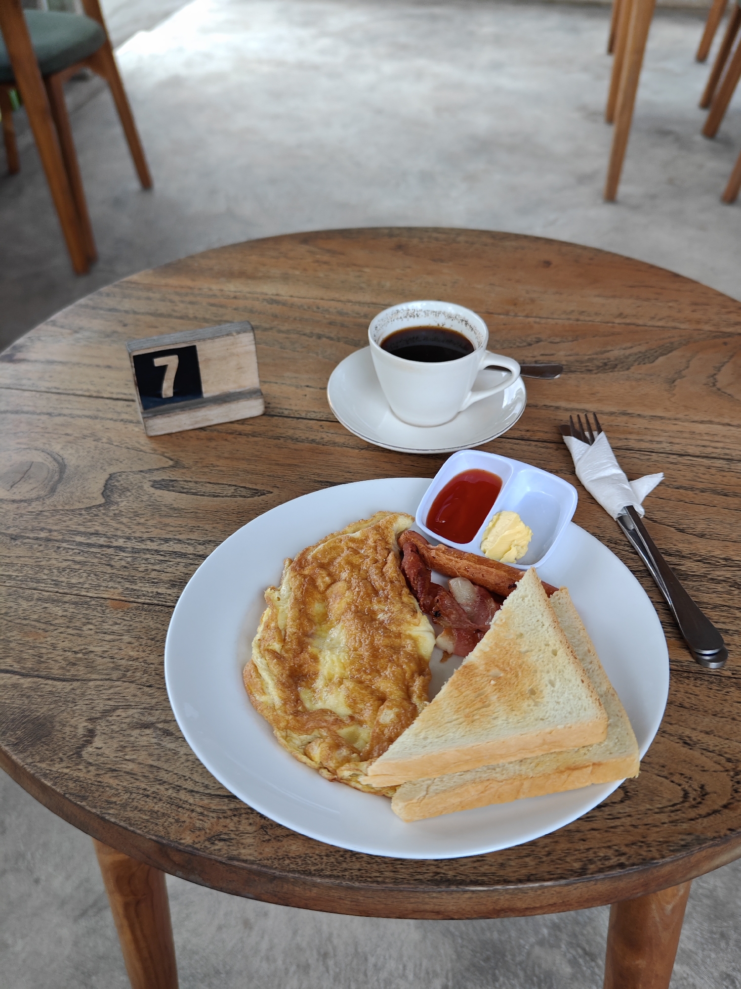A breakfast scene is captured on a wooden table in what appears to be a cafe or restaurant, potentially located in Nusa Penida, Indonesia, given the likelihood of this type of setting in the area. The table is round and has visible wood grain.
On the table is a white plate containing an omelet, two slices of toast, bacon, sausage, and a small white dish with ketchup and butter. A white cup with a saucer filled with black coffee is also on the table.
To the left of the plate, there's a wooden table number stand displaying the number "7". A fork, wrapped in a white napkin, rests to the right of the plate.
The table is positioned outdoors, with chairs visible in the blurred background, suggesting an open-air or semi-outdoor seating arrangement. The overall lighting appears to be natural daylight, illuminating the scene.