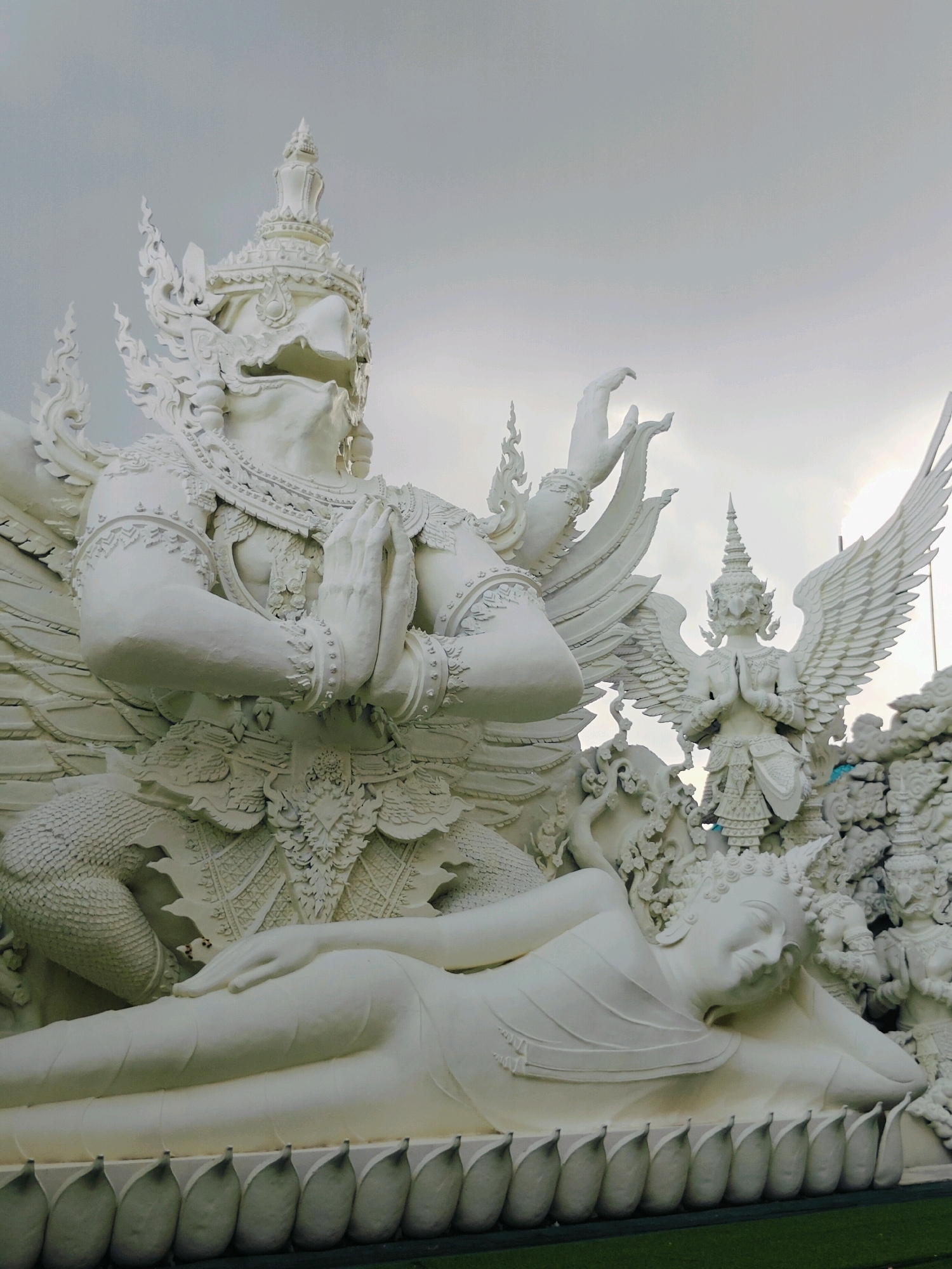 The image shows a large white sculpture of a reclining figure, possibly a Buddha, in the foreground. Behind this figure, and to the left, is a massive white sculpture of a mythical creature with a bird-like head and human-like hands clasped in prayer. This creature has intricate detailing on its armor and headdress. To the right of the reclining figure, and further in the background, is another white sculpture of a winged humanoid figure, also with hands clasped in prayer, and wearing ornate attire and a headdress. Behind these figures, more white sculptures of various beings and architectural elements are visible, creating a crowded and detailed scene. The sky is overcast and grey. The sculptures appear to be made of stucco or a similar material and are highly detailed with intricate carvings and ornamental features. The overall impression is of a significant religious or artistic monument. The location is stated as Phra Nakhon Si Ayutthaya City Municipality, Thailand.
