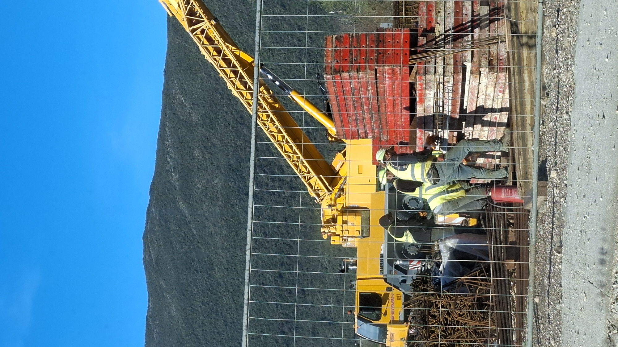 The image depicts a construction site located in Budva, Montenegro, with densely forested mountains visible in the background under a clear blue sky. In the foreground, a metal mesh construction fence partially obstructs the view. Four individuals, identified as construction workers, are present behind this fence. They wear safety vests, and at least two are wearing hard hats (one white, one green). The workers are gathered near bundles of rebar and stacks of red formwork panels. One worker is bending over, while others stand upright, appearing engaged in material handling. A large yellow Liebherr mobile crane, with "BEMAX" and "697" visible on its cab, is positioned to the left and center, its boom extending towards the upper right. A red fire extinguisher is on the ground near the workers. The ground surface consists of gravel and dirt. Sunlight casts distinct shadows to the right of the workers and materials.