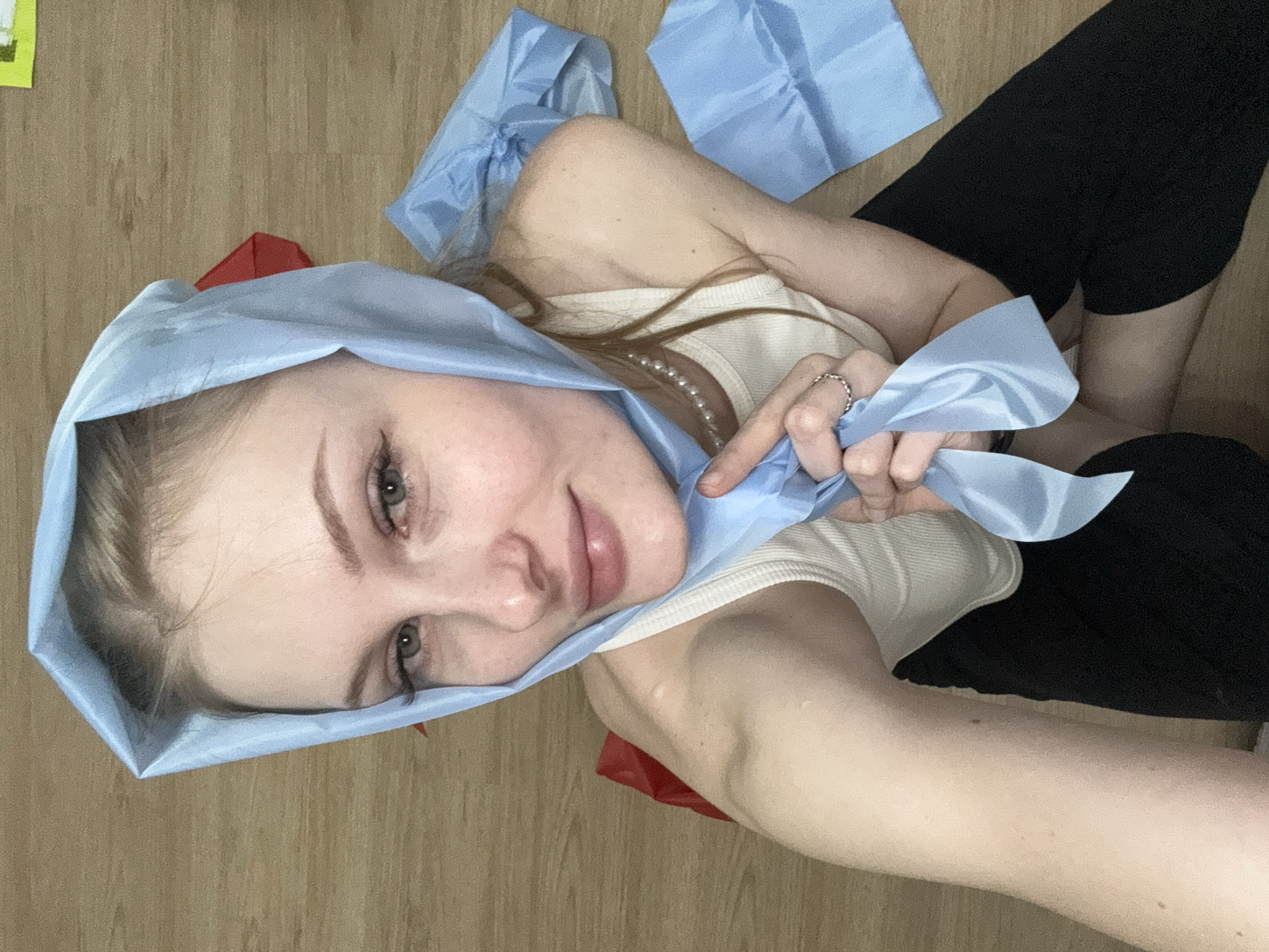 A young woman is sitting on a wooden floor, wearing a light blue headscarf and a white ribbed tank top. She has on a pearl necklace and a silver ring on her left hand. There are several pieces of blue fabric scattered around her, along with a red object partially visible under the blue scarf. The room appears to be sparsely furnished, with the wooden floor being the predominant element of the environment. The image was taken from a close-up perspective, focusing on the woman's face and upper body. No specific details about the location in the city of Perm, Russia are visible in the image.