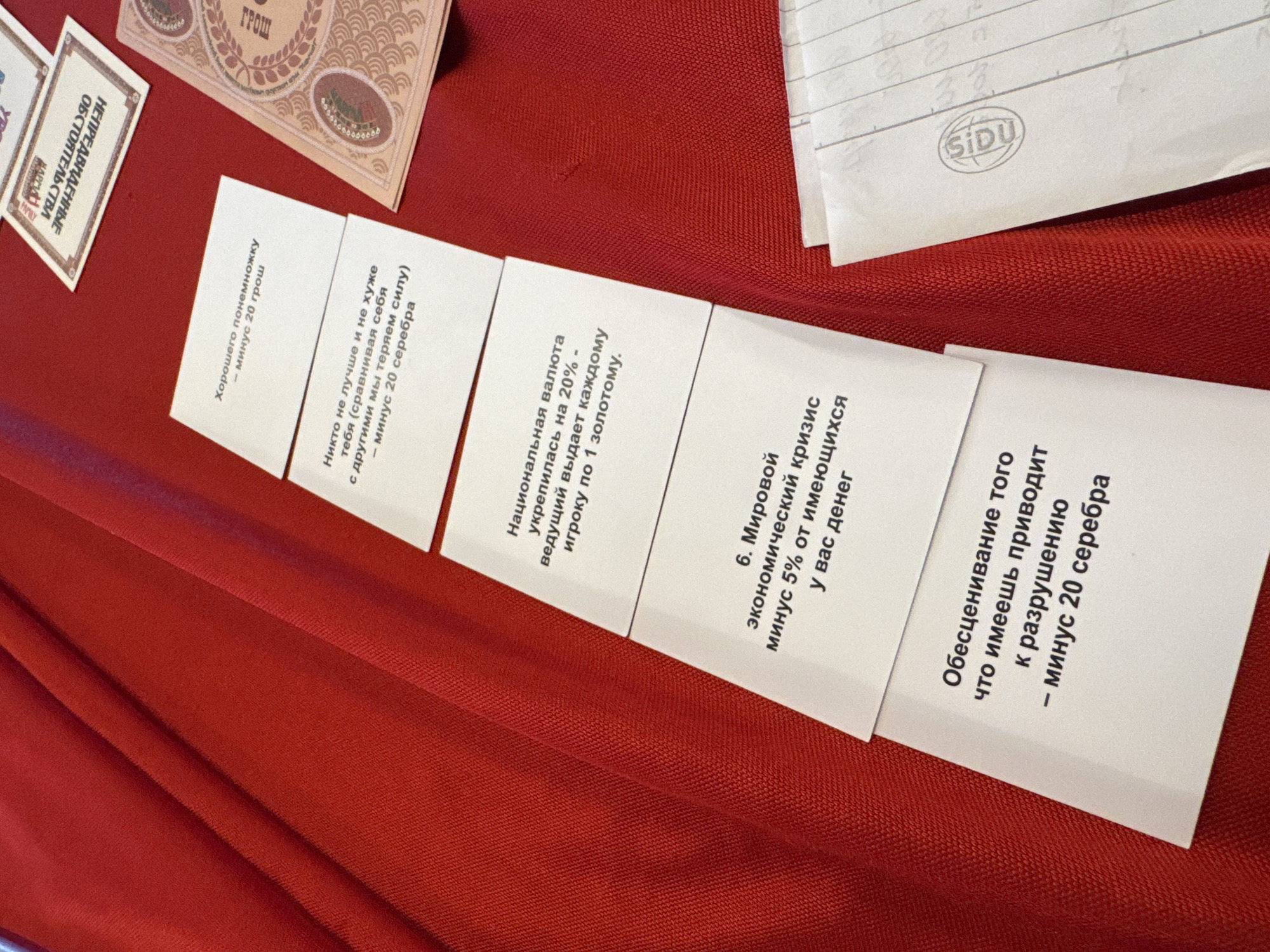The image displays an indoor scene featuring several white cards with Russian text, a larger beige card, and a white envelope, all arranged on a vibrant red, textured fabric surface. No people are visible.
The setup strongly suggests a board game or an educational simulation exercise, likely focused on economic or financial literacy themes. The cards detail various scenarios and their financial consequences, using game-specific currencies like "grosh," "silver," and "gold."
Visible text includes:
- "UNFORESEEN CIRCUMSTANCES" (on a card partially visible on the left).
- "A little good - minus 20 grosh."
- "No one is better or worse than you (comparing ourselves to others, we lose strength) - minus 20 silver."
- "National currency strengthened by 20% - the leader gives each player 1 gold."
- "6. Global economic crisis minus 5% of the money you have."
- "Devaluation of what you have leads to ruin - minus 20 silver."
A larger card in the background prominently features the word "ГРОШ" (GROSH). A white envelope with the "SIDU" logo, an Indonesian stationery brand, is visible in the upper right corner. This detail subtly ties into the provided location context of Kuta Selatan, Indonesia, despite the Russian language on the game cards. The overall mood is neutral and functional, typical of a game or educational setting.
