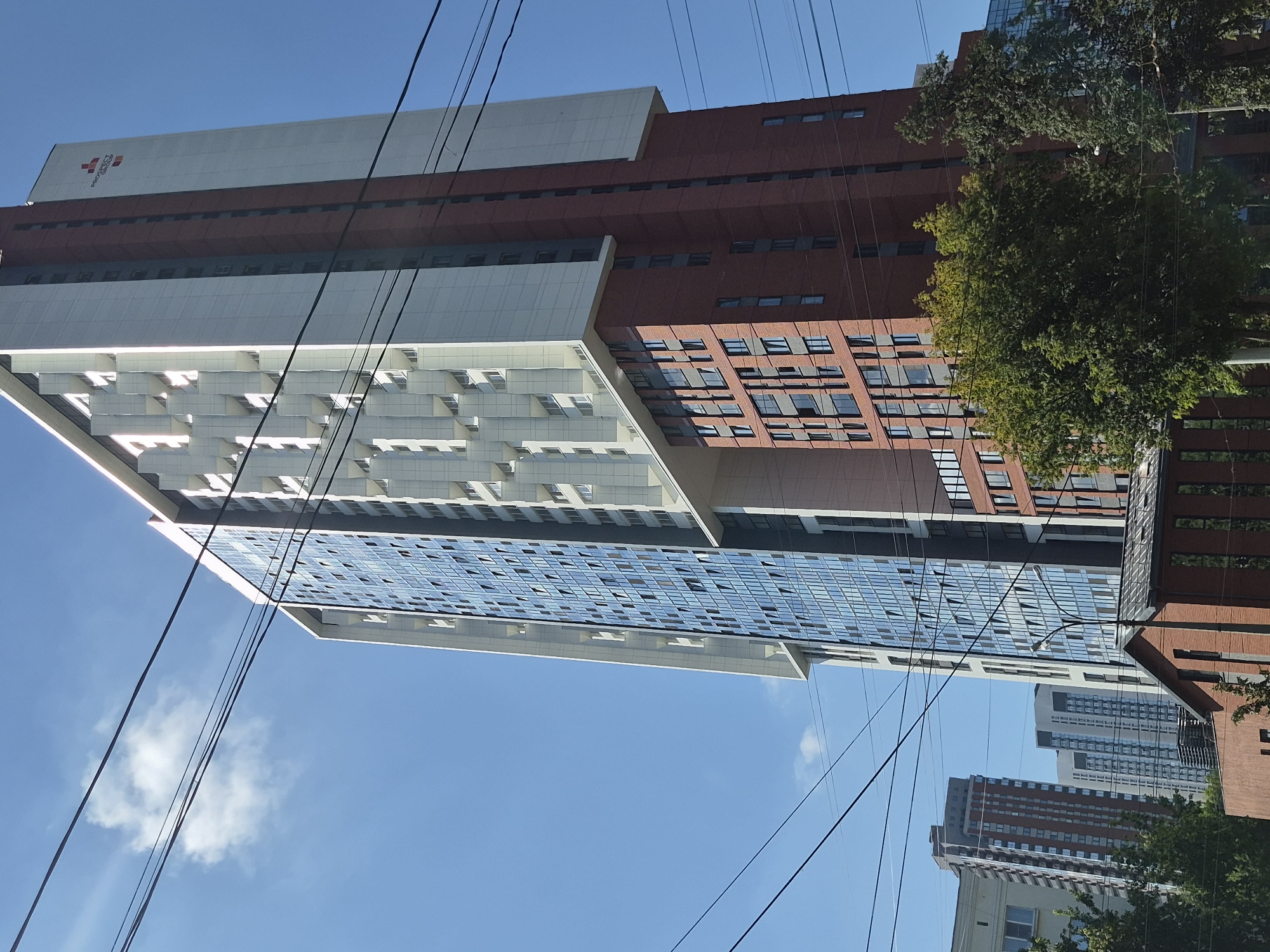The image captures a bright, sunny day in Yekaterinburg, Russia, with a clear blue sky and a few scattered clouds. The foreground is dominated by a very tall, modern residential building with a distinctive architectural design. The building features a lower section clad in red brick with numerous dark-framed windows, and an upper section that is predominantly white with a glass facade composed of many small, reflective panes. Several prominent power lines crisscross the frame, obscuring parts of the sky and the building.
Further back, a row of other multi-story buildings is visible, varying in architectural style and color, suggesting an urban environment. Green trees are interspersed throughout the scene, adding natural elements to the cityscape. One of the buildings, likely the main subject, displays a red cross logo and text that appears to read "PROSPEKT GARDEN" on its upper portion. The perspective is from below, looking up at the towering structures. No people are visible in the image. The overall impression is one of a contemporary urban landscape under a clear sky.