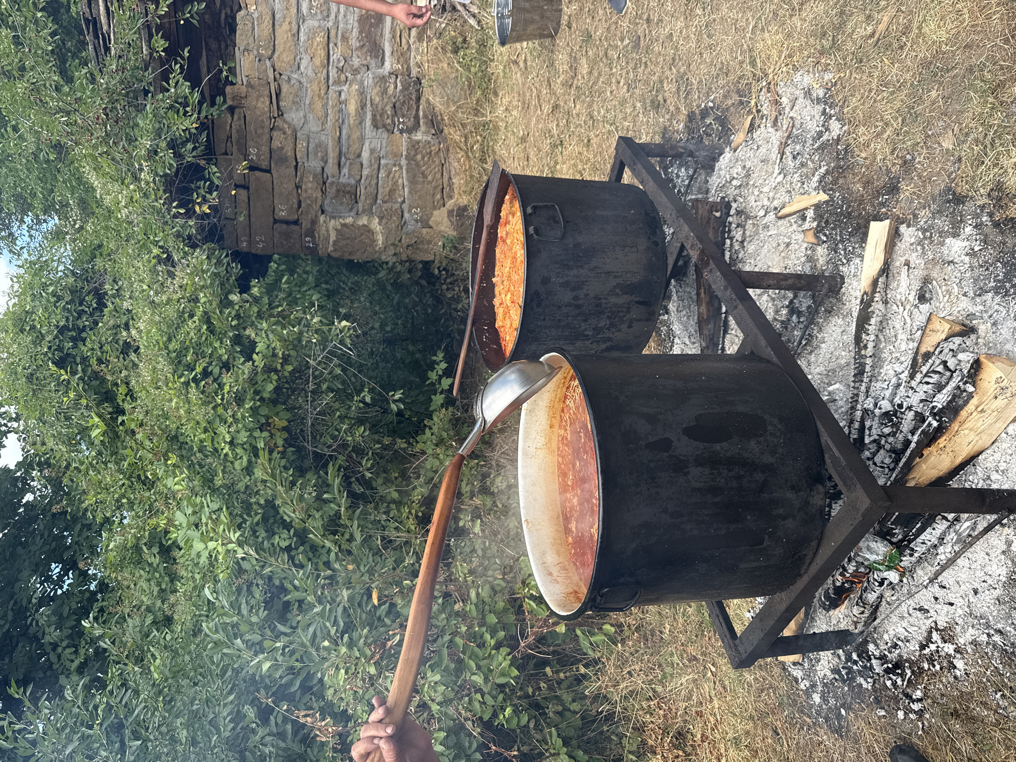 The image depicts an outdoor cooking scene in Manaselska Reka, Bulgaria, with two large black pots suspended over a wood fire. The left pot contains a reddish stew, and a ladle is being used to stir it. The right pot holds a orange-colored stew. In the background, a stone structure with a weathered roof is visible, partially obscured by dense green foliage. The ground is covered with dry grass and the remnants of a fire, including ashes and scattered logs. A hand is visible on the ladle, and another person's arm and legs are partially visible on the right side of the frame. A metal bucket sits near the stone structure. The scene suggests a communal or outdoor cooking event.