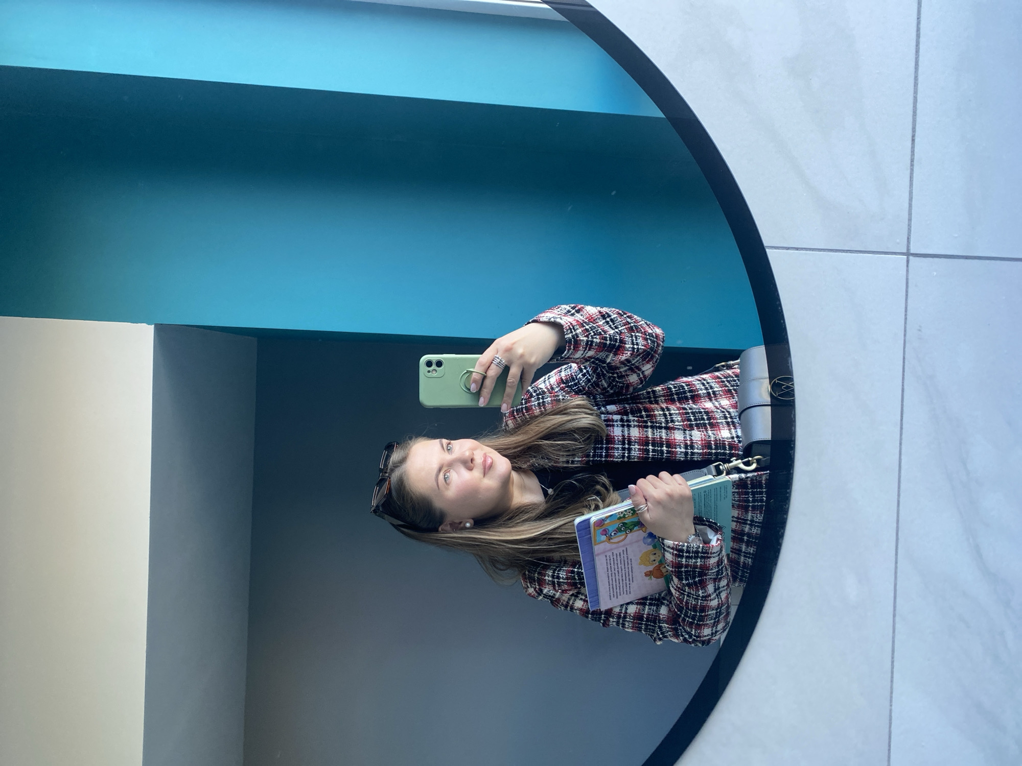A woman is taking a selfie in a circular mirror. She is wearing a plaid jacket and holding a book and a cell phone with a green case. Her sunglasses are perched on her head. The background appears to be a hallway with walls painted in shades of white and blue. The location is identifiable as the Central Children's Library in Izhevsk, Russia, based on the distinctive architectural style and color palette of the interior visible in the reflection.