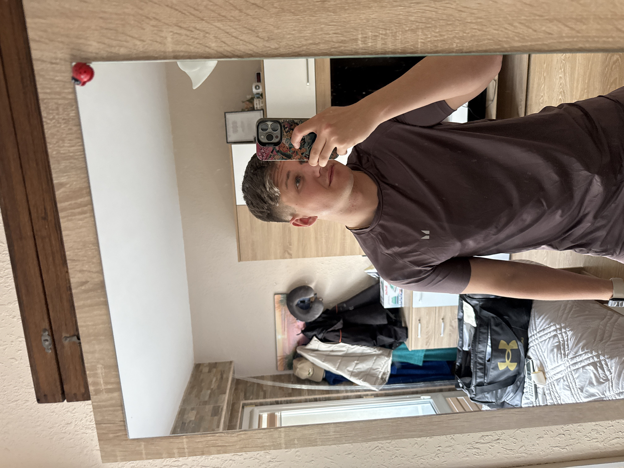 A person is taking a selfie in a mirror. The person has short brown hair and is wearing a brown shirt. The mirror is framed with a light-colored wood. In the reflection, there is a bed with a white quilt, a black Under Armour bag, and a window. Also visible are a wall with wood-like tiles, clothing, a framed picture, and cabinets. The location cannot be determined to be Burgas, Bulgaria from the visual information provided.