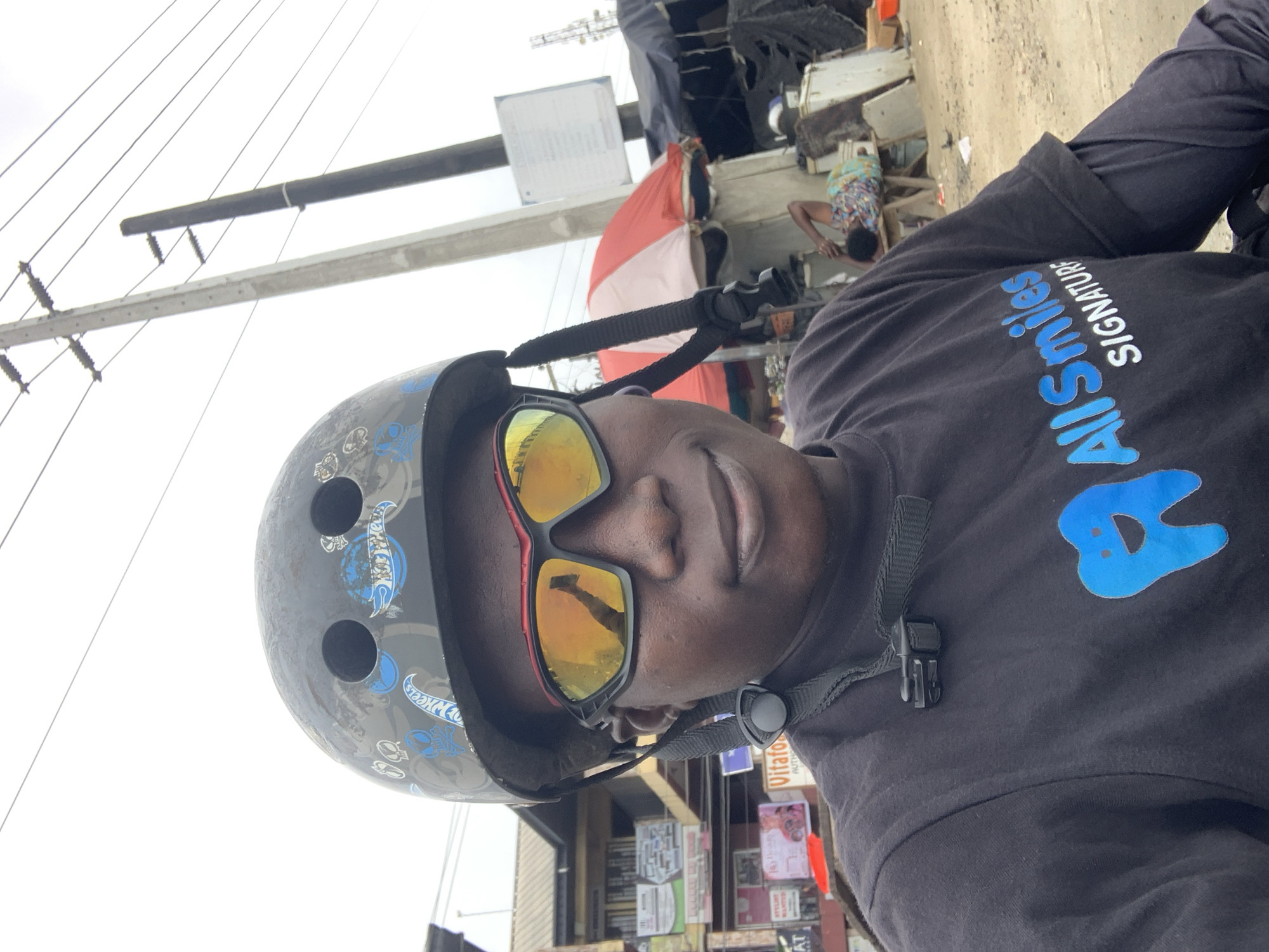 This is a frontal, eye-level selfie taken outdoors in what appears to be a bustling urban environment in Lagos, Nigeria. The subject, a person with dark skin, is wearing a black t-shirt with a blue tooth logo and the text "AllSmiles SIGNATURE." They are also wearing a black helmet adorned with "Hot Wheels" decals and orange-tinted sunglasses. The background features utility poles with electrical wires, buildings with signage, red and white umbrellas, and other structures suggesting a marketplace or informal settlement. In the mid-ground, a person is lying down on a wooden bench beneath an umbrella. The ground is dusty and unpaved. The overall atmosphere is lively and somewhat chaotic, typical of street scenes in Lagos.