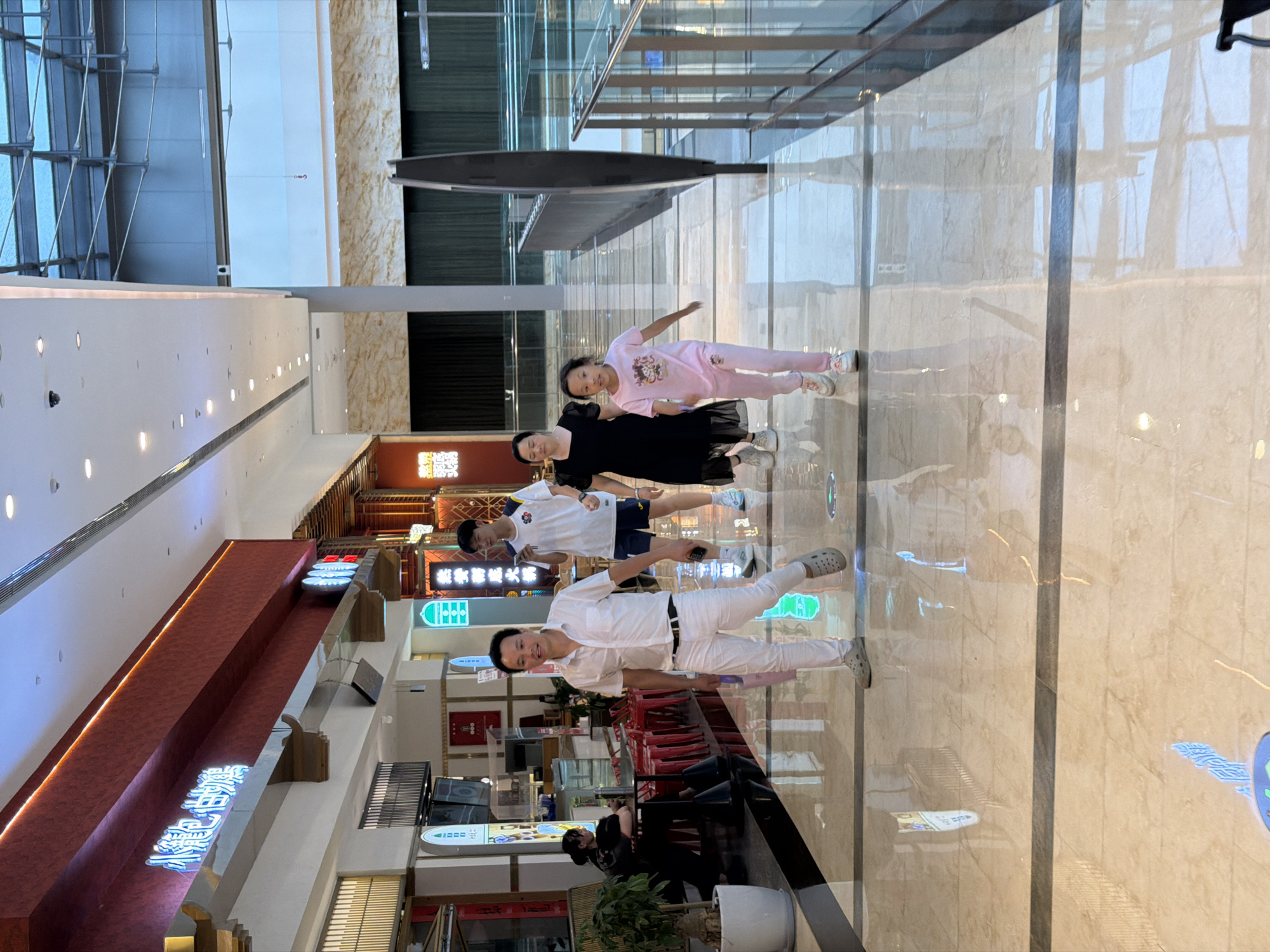 The image depicts an indoor walkway in what appears to be a shopping mall or commercial complex in Yuzhong District, China. The floor is polished marble with dark grey inlaid strips, reflecting the overhead lighting and architectural elements.
In the foreground, a family is walking towards the camera. A man in a white short-sleeved collared shirt and white pants, wearing grey slip-on shoes, is in the lead. To his right, a woman in a black dress and white sneakers follows. A young girl in pink pants and a pink t-shirt, also wearing white sneakers, walks beside the woman, slightly ahead.
Further back, a young man in a white t-shirt and blue shorts is visible, holding a phone. To the left, in the background, a woman is seated at a table with red chairs, facing away from the camera. Various shop fronts and signage are visible along the walkway, including some with blue neon lettering. The ceiling features modern lighting and architectural beams.