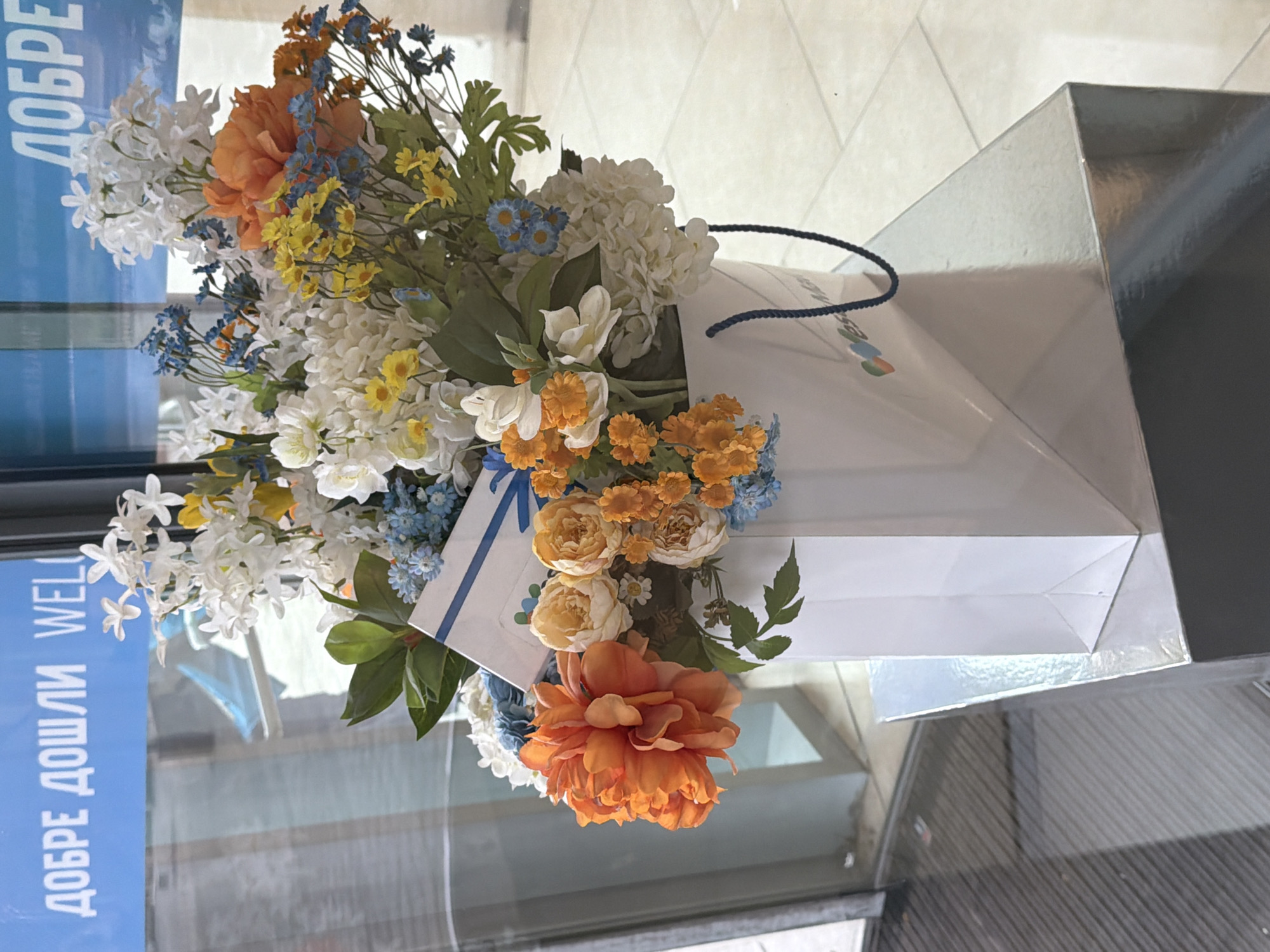A floral bouquet is arranged in a white shopping bag, resting on what appears to be a reflective surface. The flowers are a mix of white, orange, and blue hues, suggesting a vibrant artificial or fresh arrangement. The shopping bag has dark blue handles. The scene is viewed through a glass window or door, where the reflection of a sign written in Cyrillic script is visible. The location is in the city of Unknown, Unknown.