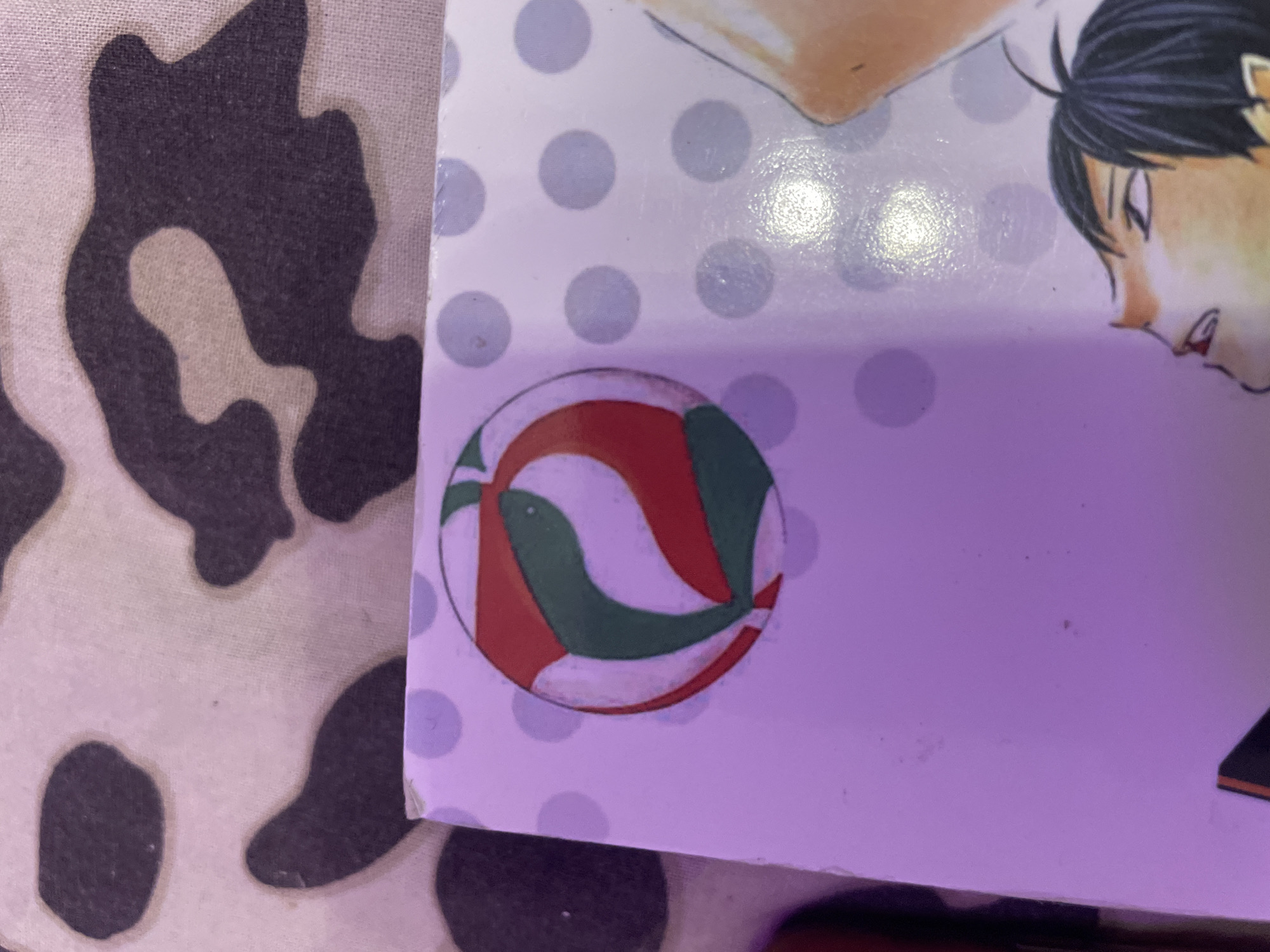 The image is a close-up, eye-level, indoor shot of a manga book cover. The cover features a volleyball with red and green markings on a light purple background with scattered light grey polka dots. The upper portion of the image shows a textured fabric with irregular dark brown and light beige patterns, possibly a blanket or clothing. The bottom right of the cover shows a partial illustration of an anime-style character's face looking upwards, with dark hair and yellow-toned skin. The scene is illuminated by a purple light source, casting a purplish hue on the book cover. The image does not contain any information about the city of Irinovka, Russia.