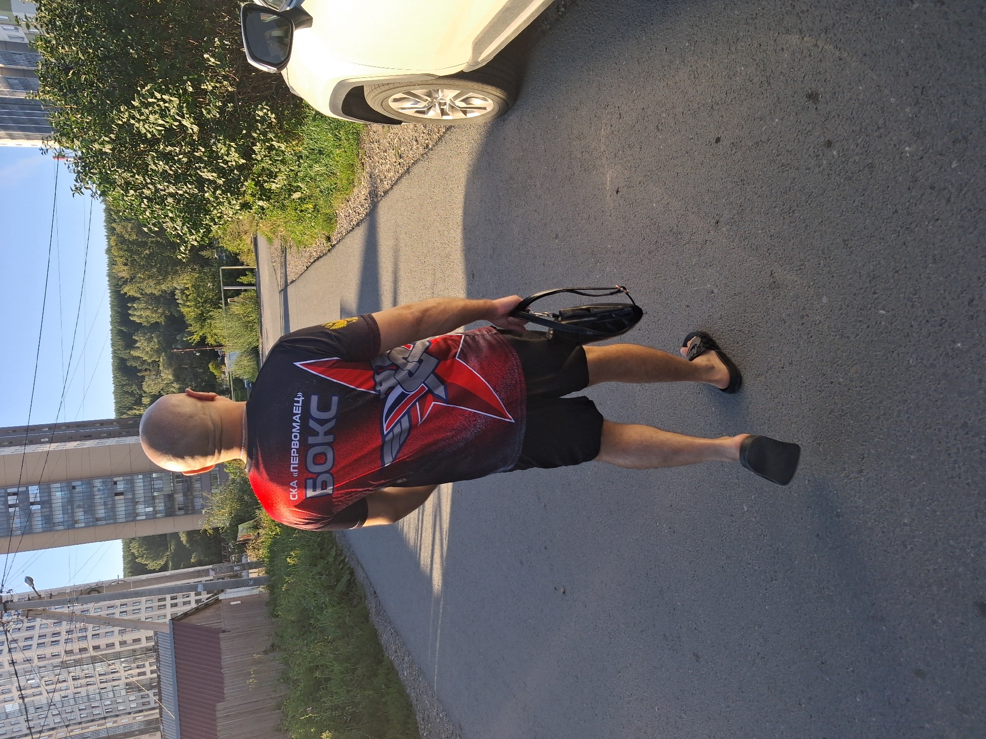 An adult male with a shaved head walks away from the camera down an asphalt road lined with grass and trees. He is wearing a black t-shirt with a red and white graphic design featuring the word "БОКС" (BOX) and a stylized winged emblem. He also wears black shorts and black flip-flops. He carries a black bag with a strap in his right hand. To the right of the road is the side of a white SUV. In the background, there are tall, modern buildings, some with many windows. A wooden structure with a red roof is visible to the left, and a utility pole with wires stands nearby. The sky is clear and blue, indicating daytime. The environment appears to be a mixed urban and semi-rural area, possibly on the outskirts of a city. The presence of tall buildings suggests an urban setting, while the vegetation and road indicate it's not the city center. This scene is consistent with a location within the Akademgorodok district of Novosibirsk, Russia, a science and education center known for its modernist architecture interspersed with green spaces.