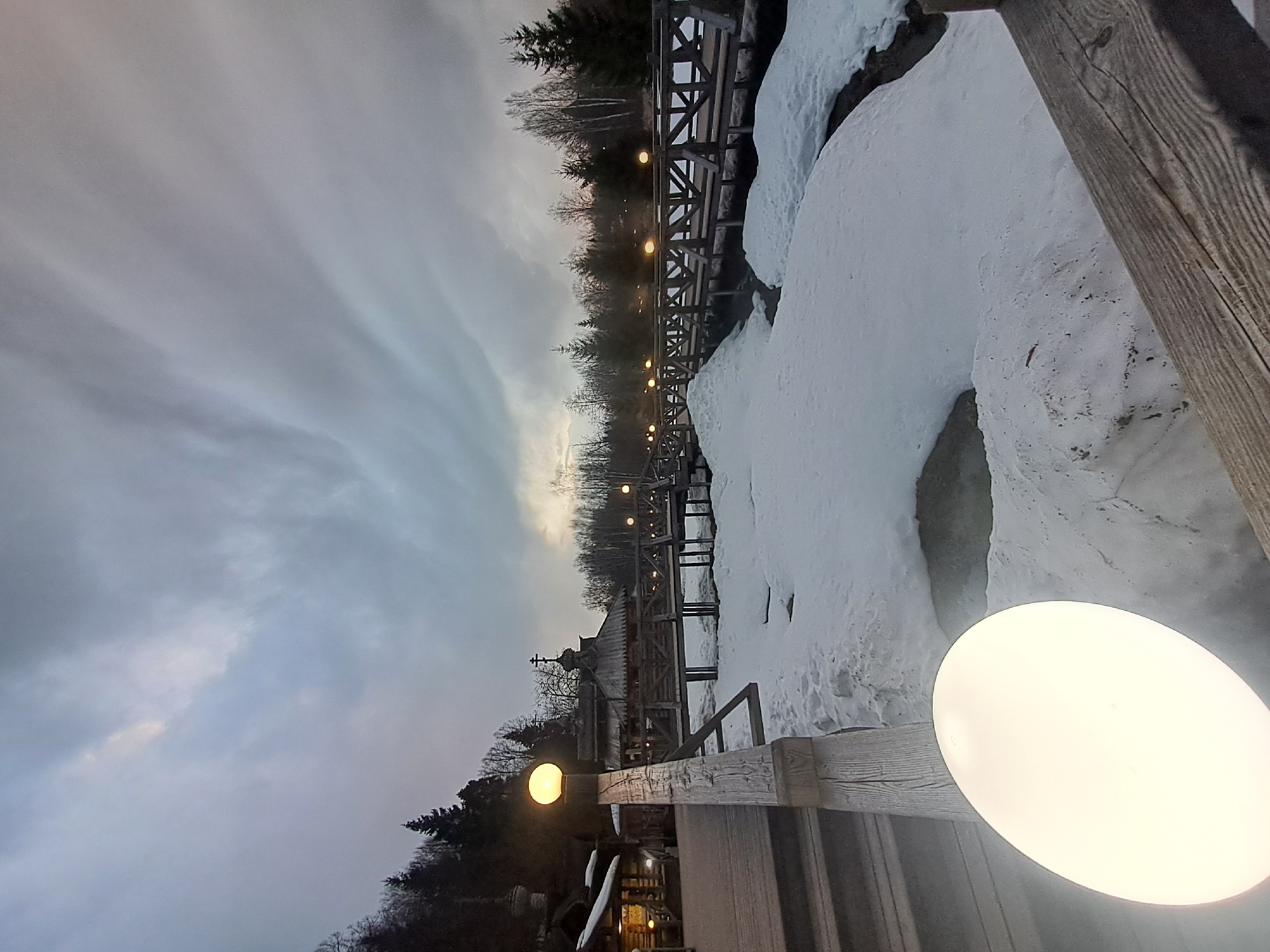 The image depicts a landscape in Vzglyadnevo, Russia, featuring a wooden boardwalk or bridge covered in snow. The scene is dominated by overcast skies. A traditional Russian wooden church with a cross on top is visible in the background, partly obscured by trees. The boardwalk is lined with illuminated lampposts. Snow covers the ground on either side of the boardwalk. A portion of a wooden railing or structure is prominently featured in the foreground, partially obscuring a lamppost.