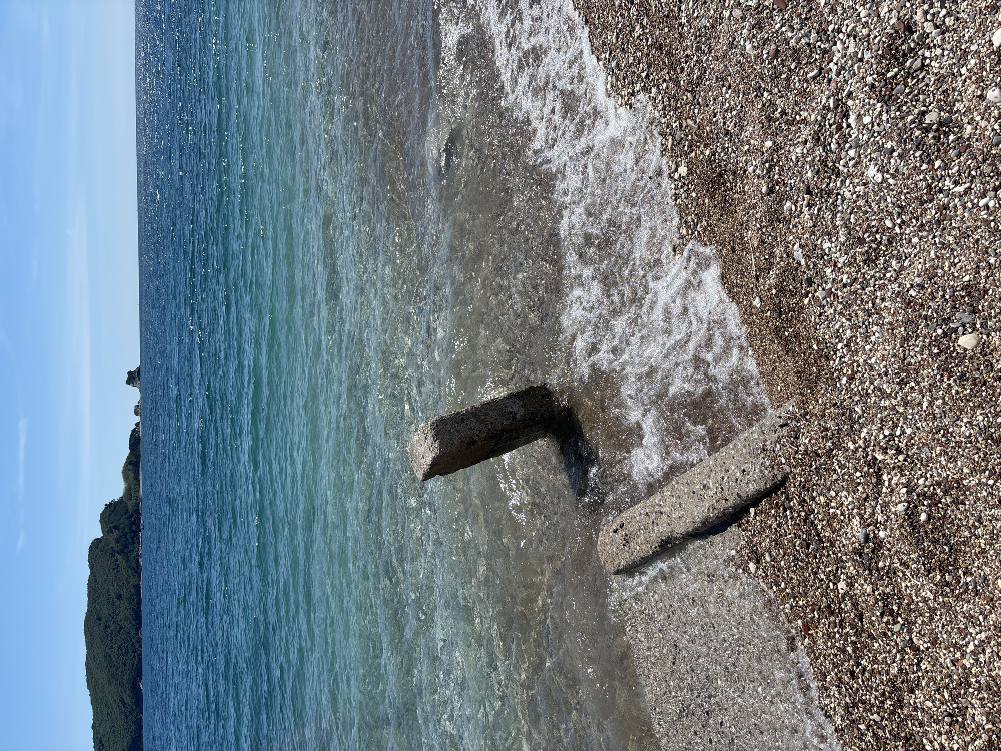 The image shows a beach scene in Budva, Montenegro. The foreground consists of a pebbled beach meeting the turquoise water of the Adriatic Sea. Two concrete structures or remnants of a structure are partially submerged in the water, close to the shoreline. A forested coastline is visible in the background under a blue sky. The scene suggests a calm day at the beach, with clear water gently washing over the pebbles and the concrete structures.