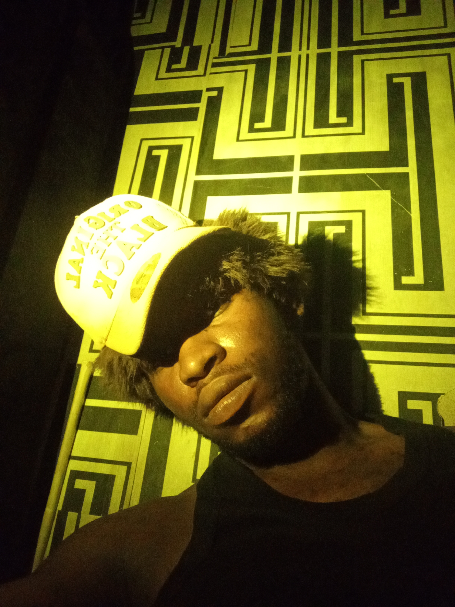 A low-angle, close-up selfie of a young Black male wearing a white baseball cap with yellow lettering and a black tank top is captured in what appears to be a poorly lit indoor setting. The background features a geometric pattern in yellow and black, possibly wallpaper or a mural. The man's face is partially obscured by shadow from his cap, but his lips are visible and appear slightly parted. His skin is dark, and he has a beard. The lighting casts a strong yellow hue over the scene. The overall impression is that of a candid, informal photograph, likely taken at night or in a dimly lit area within a residential or entertainment space in Lokoja, Nigeria.