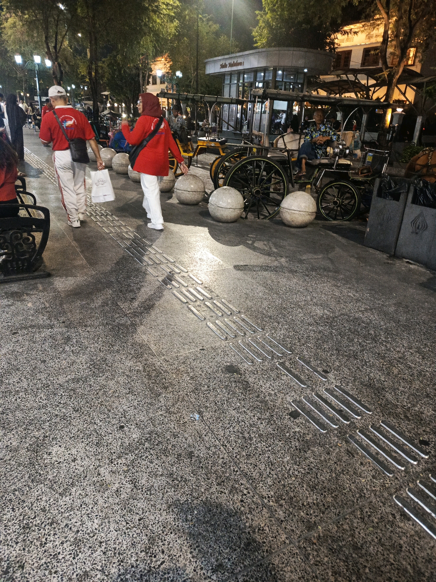The image depicts a nighttime scene on a pedestrian walkway in Malioboro Street, Yogyakarta, Indonesia. Two individuals, a man wearing a white cap and a woman in a red hijab, are walking away from the camera. The man is wearing a red t-shirt with a logo and white pants, carrying a white paper bag in his left hand and a black crossbody bag. The woman is wearing a red long-sleeved shirt and white pants, also with a black crossbody bag. In the background, several horse-drawn carriages, known as "andong," are parked, some with passengers. A man in a patterned shirt is seated in one of the andongs, appearing to be a driver or attendant. Streetlights illuminate the walkway, casting shadows from the trees and people. The walkway is paved with textured tiles, including tactile paving for the visually impaired. Buildings with illuminated windows are visible in the background, suggesting shops or residences. The overall atmosphere is that of a bustling evening street scene.