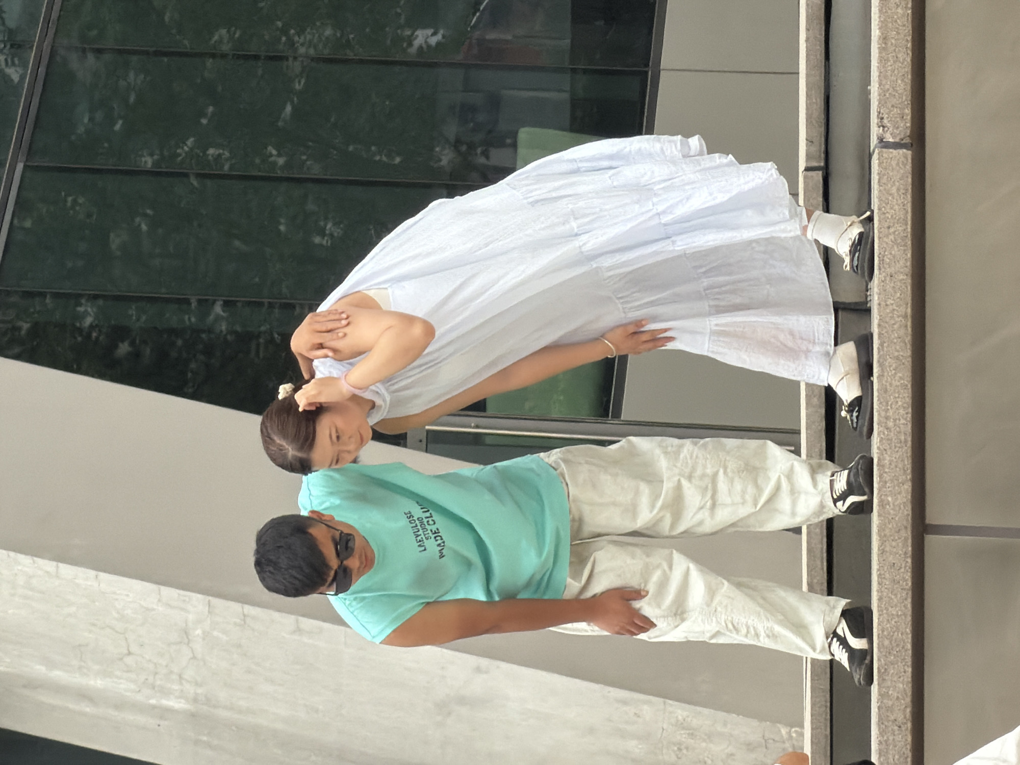 Two individuals are standing on a tiered concrete structure, possibly in a public space in Yuzhong District, China. A man on the left, wearing a turquoise sleeveless t-shirt with text reading "LAEYULOSE STUDIO MADE CLUB" and off-white baggy pants, leans forward with his left hand on his knee. He is also wearing black and white sneakers and sunglasses. A woman on the right, dressed in a flowing light blue tiered dress and white socks, leans forward with her right arm around the man's shoulders and her left hand resting on her hip. She is wearing black and white flat shoes. The background features a modern building with dark, reflective glass panels.