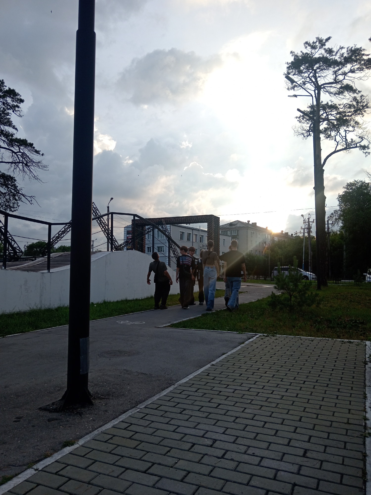A group of five individuals, appearing to be adult males of varying ages and builds, are walking away from the camera on a paved sidewalk. The individual in the lead wears a dark t-shirt and blue jeans. The second person from the front is wearing a lighter colored t-shirt and dark pants. The third person is wearing a striped shirt and jeans. The fourth person is wearing a t-shirt and dark pants. The last person in the group is wearing a dark t-shirt and jeans. They are walking on a sidewalk that transitions from asphalt to interlocking pavers. To the left of the sidewalk is a grassy area and a low white wall, beyond which are metal structures that appear to be part of a sports or recreational facility. In the background, there are multi-story buildings and trees. The sky is cloudy with the sun breaking through, creating a bright, hazy effect. A tall, dark lamppost is prominently positioned in the foreground on the left side of the image, and another tree with a street lamp attached to it is visible on the right side. The image appears to have been taken in the city of Belogorsk, Russia, given the architectural style of the buildings and the general environment.