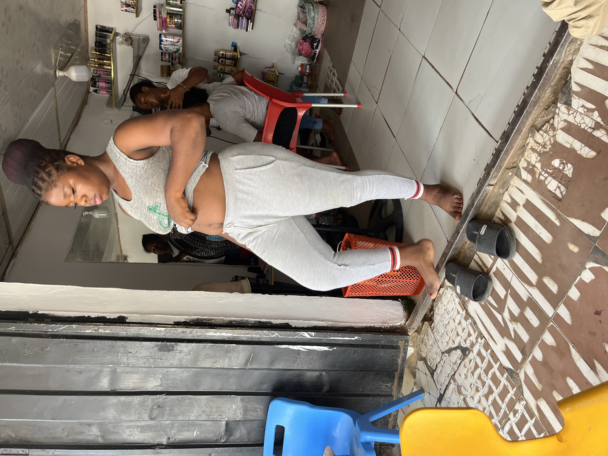 The image, rotated 90 degrees clockwise, depicts an indoor scene, likely a small beauty salon or hair braiding shop. In the foreground, a woman stands facing left, wearing a grey sleeveless top and light grey leggings. Her hair is styled in dark braids pulled into an updo. She appears to be adjusting her top or leggings around her waist, looking downwards with a neutral expression.
In the mid-ground, two other individuals are present. One person, also in a grey top, is seated on a red plastic chair, receiving attention to their hair from a third person whose head is visible behind them. The background features shelves stocked with various beauty products, including bottles, containers, and accessories. A white bottle stands on one of the shelves. A black fan is partially visible behind the standing woman.
The floor is tiled with two distinct patterns: a white and brown geometric design on the right, and a lighter, mostly white pattern with visible cracks towards the left. A raised threshold separates these two tiled sections. Near the standing woman's feet, a pair of black slide sandals, marked with the text "OOFOS," rests on the floor. Parts of a blue and a yellow plastic chair are visible in the lower foreground. The far left of the image shows a dark, paneled structure, possibly a wall or entrance. The lighting suggests an indoor setting, with no specific time of day discernible. The scene captures a typical daily activity within a local business environment, consistent with locations like Abuja, Nigeria.