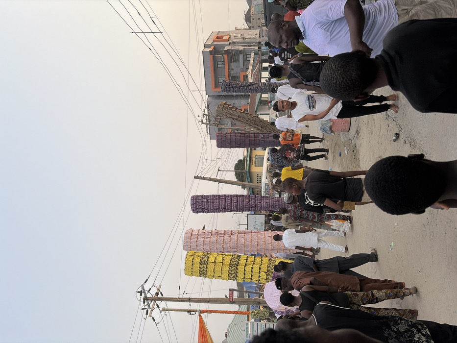 An outdoor scene displays a crowd of people lining a street next to vertical structures. These are decorated with wrapped materials of yellow, pink, and purple patterns. The crowd is diverse in attire, with some wearing white shirts. Overhead, power lines crisscross the sky near buildings. The street appears somewhat dusty, with a mix of light and shadows.