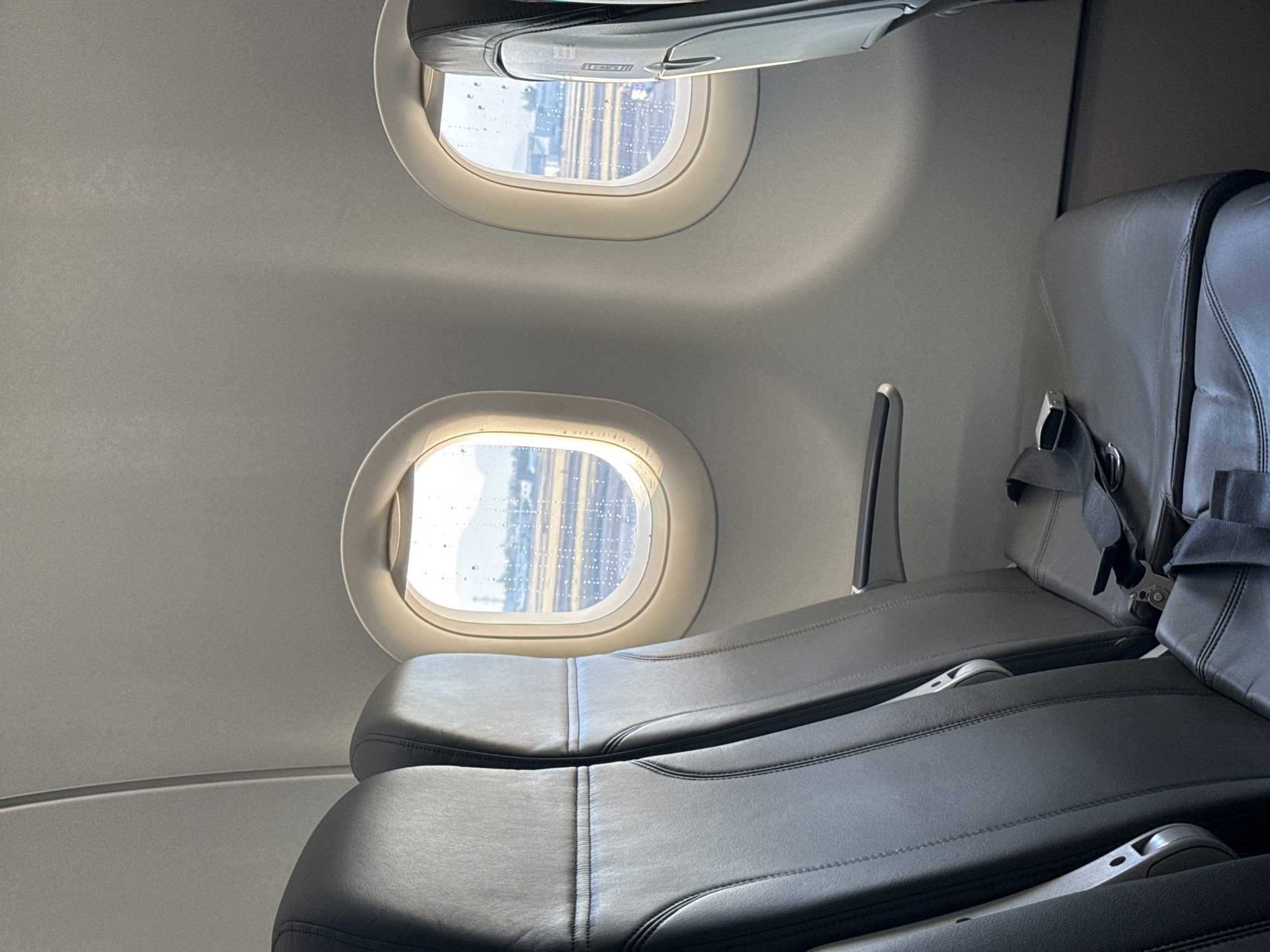 The image shows the interior of an airplane, likely during boarding or taxiing, based on the view outside the windows. Two oval-shaped windows are visible on the left side of the frame, revealing a cityscape with tall buildings outside, likely taken in Mexico City, Mexico. The windows are covered with rain droplets. To the right of the windows, two dark gray leather passenger seats are visible, featuring adjustable headrests and seatbelts. The cabin walls are light gray. The seating configuration appears to be two seats per row.