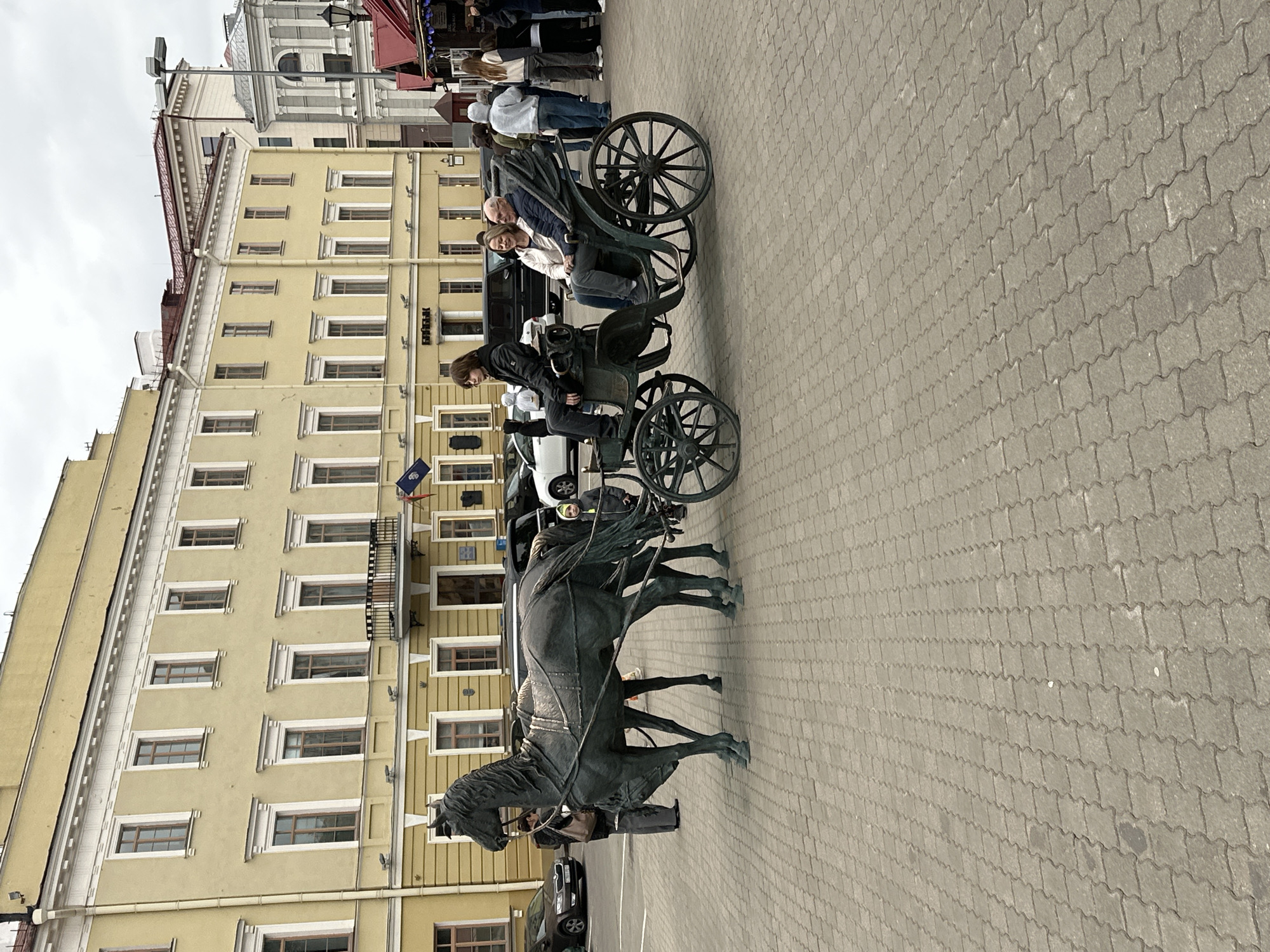 The image captures a street scene in Minsk, Belarus, featuring a horse-drawn carriage sculpture. A person is sitting on the driver's seat of the sculpture. Another pair of people is sitting in the passenger seats. In the background, a yellow building stands tall, with numerous windows visible on its facade. People can be seen in the background as well. The ground is covered with interlocking pavement stones. Several cars are parked nearby. The weather appears overcast.