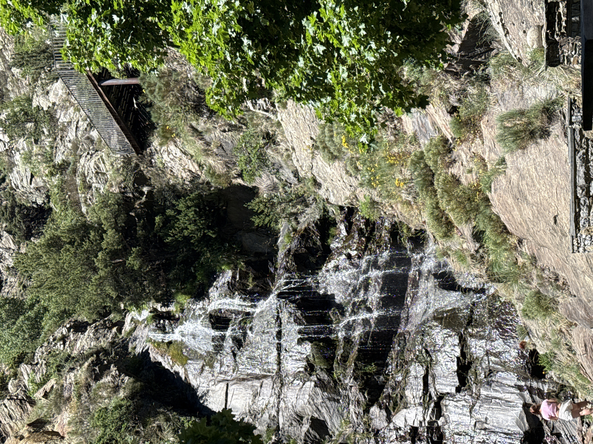 The image depicts a steep, rocky mountainside with a cascading waterfall. A metal footbridge, likely part of a hiking trail, is visible on the upper left, leading over a crevice. Lush green foliage, including trees and smaller plants, grows on the rocks, especially in the shaded areas. The waterfall tumbles down a dark, wet rock face, creating streaks of white water that sparkle in the sunlight. In the lower right, a person wearing a pink shirt and white shorts is standing, observing the waterfall. The scene appears to be set during the daytime, with bright sunlight illuminating the rocks and water. The overall impression is one of a natural, rugged landscape, possibly a popular spot for outdoor recreation.