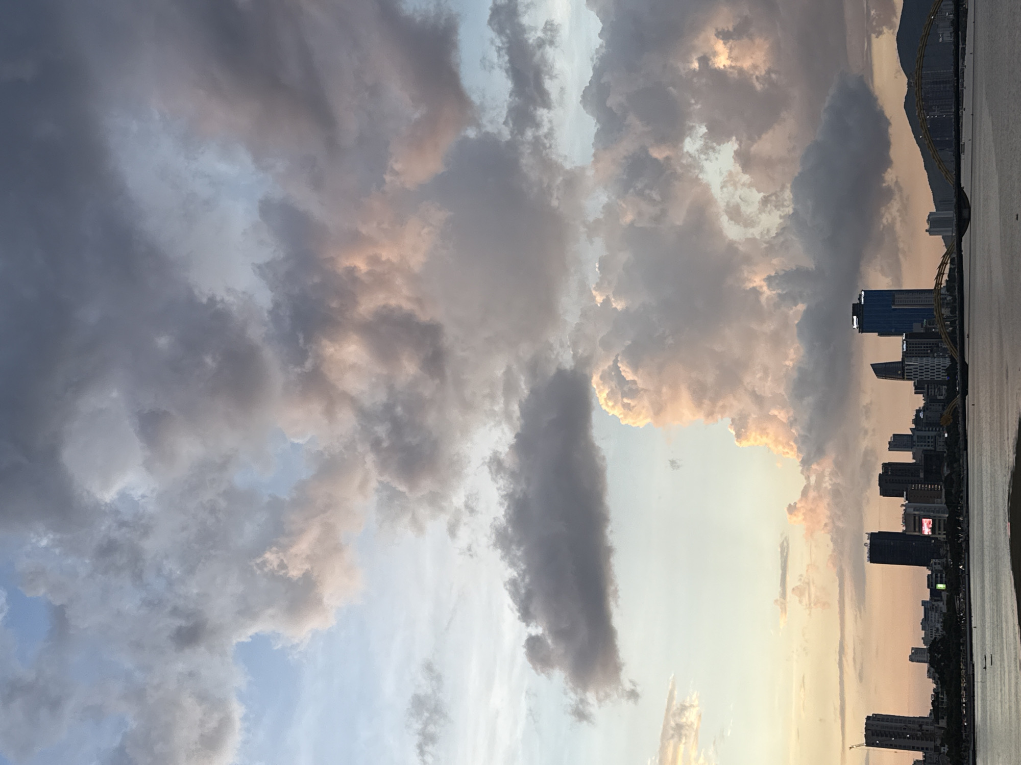 The image captures a panoramic view of the Da Nang cityscape at sunset. The foreground features a body of water, likely the Han River, with a distant shoreline lined with numerous tall buildings and skyscrapers, characteristic of a modern urban center. A distinctive arched bridge, possibly the Dragon Bridge, spans the river, its yellow structure silhouetted against the sky. In the background, mountains rise, providing a natural backdrop to the city. The sky is dominated by dramatic clouds, illuminated by the warm hues of the setting sun, with a mix of soft pinks, oranges, and grays. The overall scene depicts a vibrant coastal city at dusk, with a blend of architectural marvels and natural beauty.