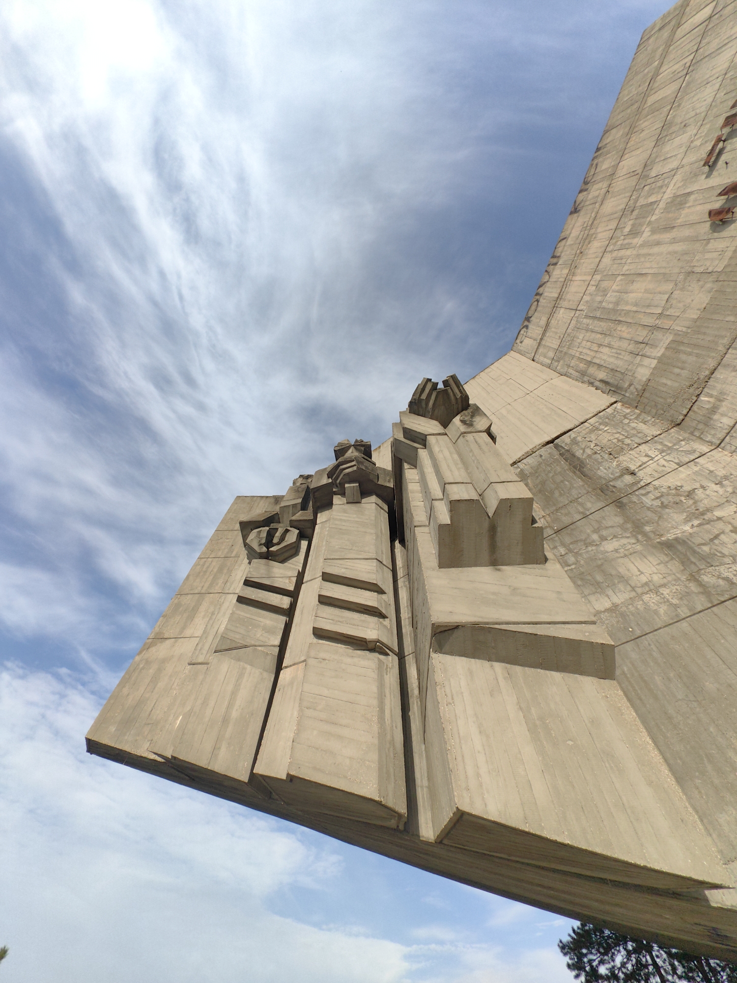 The image shows a low-angle view of the Monument of the Bulgarian-Soviet Friendship in Varna, Bulgaria. The large concrete structure dominates the frame, with its brutalist architectural style clearly visible. The monument is made up of large, angular segments and has textured surfaces. The sky is visible in the background with clouds scattered throughout the blue sky. Some greenery, likely trees, can be seen at the bottom right corner of the frame. The monument appears weathered, and its colour is mostly gray.