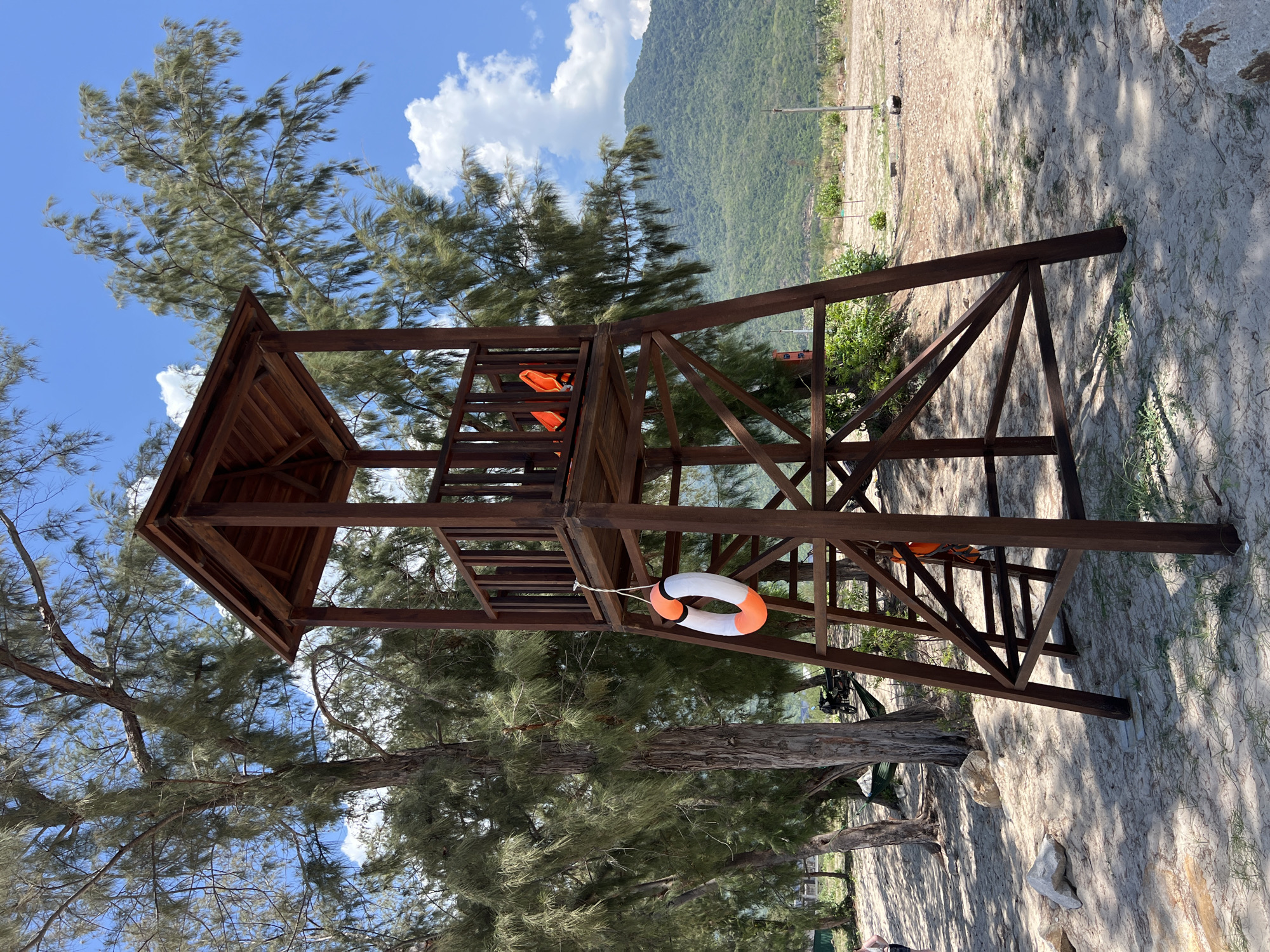 A wooden lifeguard tower stands on a sandy beach in Cong Hai, Vietnam. The tower has a square platform with railings and a roof. A lifebuoy with orange and white stripes hangs from the side of the tower. Two orange life vests are placed on the platform. Trees with long, thin leaves surround the tower. In the background, a lush green mountain rises against a blue sky with scattered white clouds. The beach is sparsely populated with rocks and vegetation.