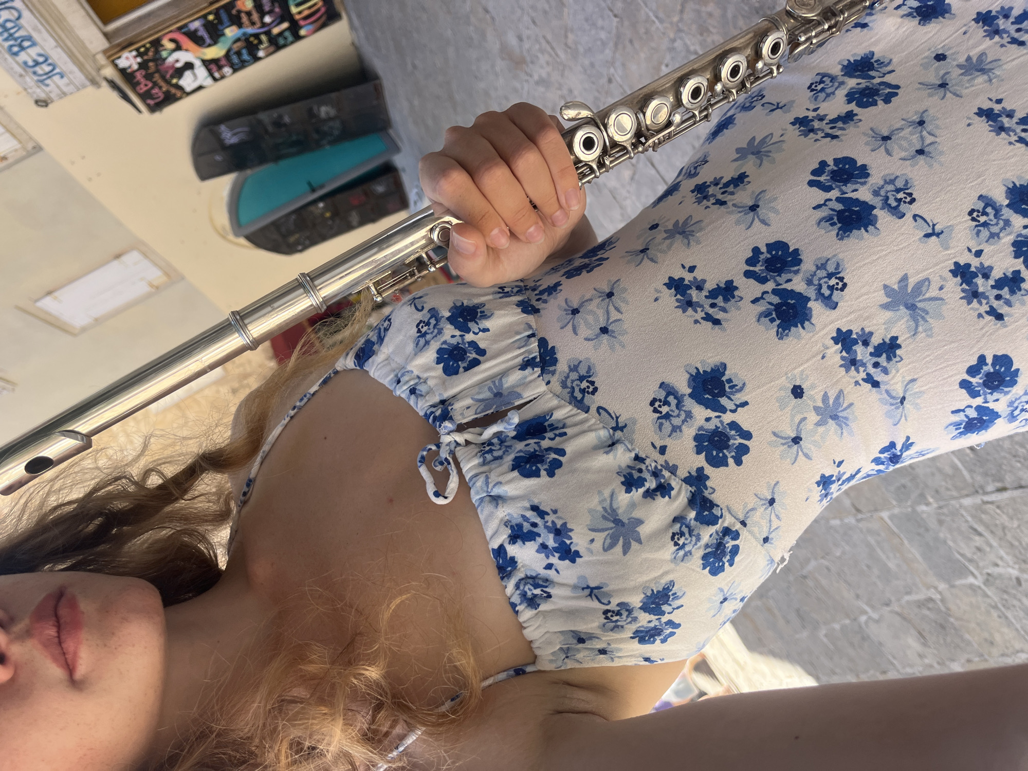 A woman holding a flute is positioned facing the camera, possibly in the city of Budva, Montenegro. She is wearing a white sundress with blue floral patterns. The flute is silver, and her hands are positioned to play it. In the background is a building with a sign saying "ICE BABY".