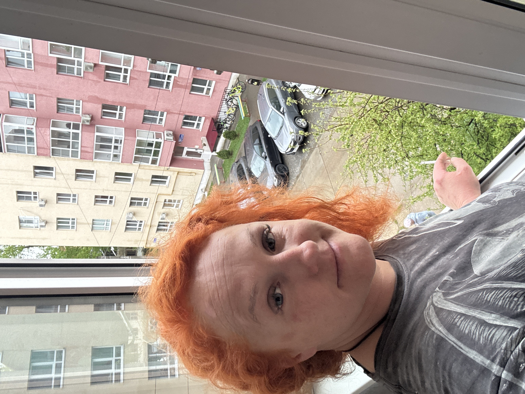 A woman with orange hair is positioned in front of a window. She is wearing a gray t-shirt with a design. Outside the window is a view of a multi-story apartment building with a pink and tan facade. Cars are parked on the street below, and trees with green foliage are visible. The location is in the city of Chaikovsky, Russia.
