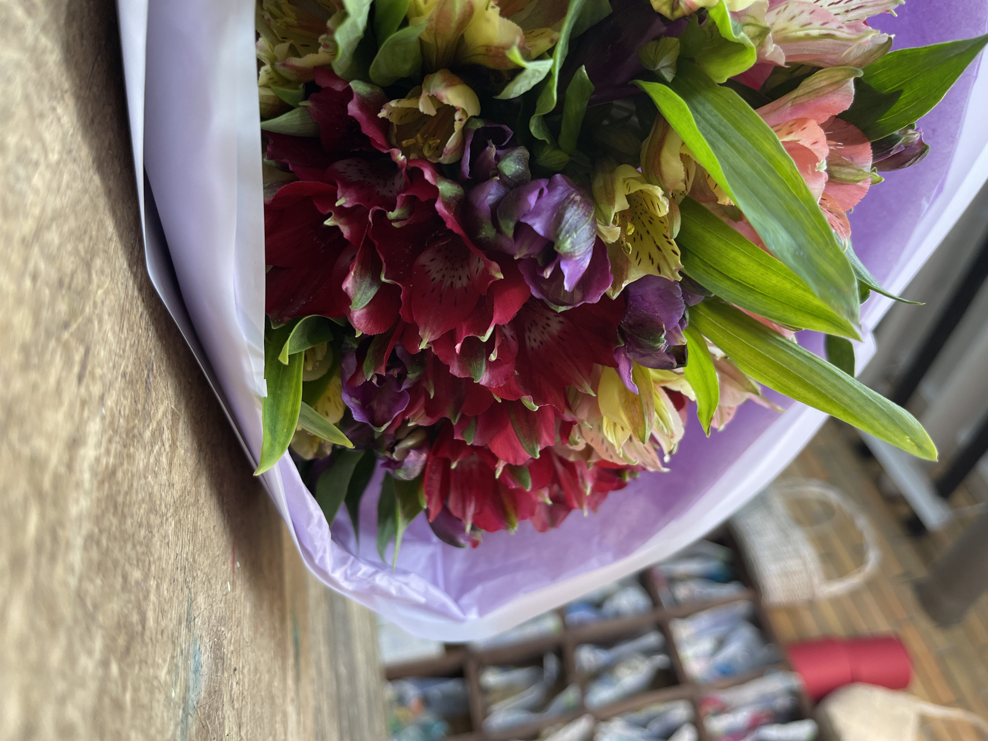 A close-up, slightly angled shot shows a bouquet of alstroemeria flowers wrapped in iridescent purple paper, lying on a wooden surface. The flowers are a mix of red, pink, yellow, and purple, with green leaves peeking out. The background is blurred, showing a glimpse of a brick wall, shelves filled with items, and a white woven basket. There are no people or specific landmarks identifiable to pinpoint the location in Kommunarka, Russia.