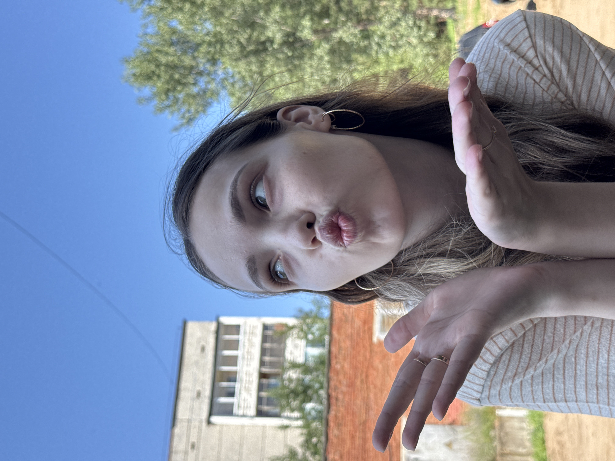 A young woman is captured in a close-up outdoor shot, outdoors. She is facing left, with her head tilted upwards and her lips puckered as if blowing a kiss. Her eyes are looking upwards and to the left. Her right hand is extended towards the viewer, palm open, while her left hand is also extended, fingers splayed. She is wearing a striped shirt and hoop earrings. The background includes a brick building, an apartment building, and lush green trees, suggesting an urban or suburban setting. The sky is a clear blue. The scene appears to be set in Noviy, Russia, given the architectural style of the buildings.