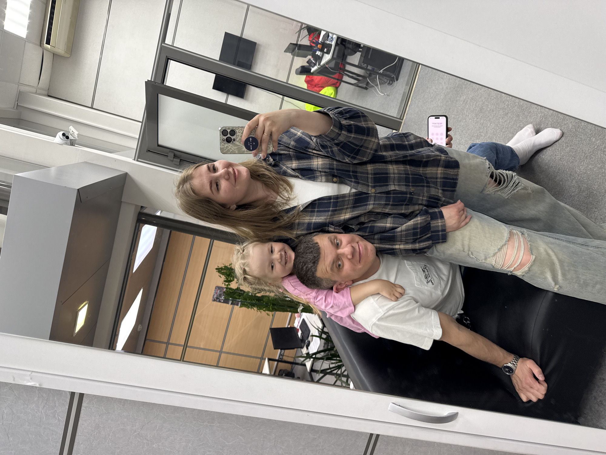 The image shows a family of three reflected in a mirror. A young woman is taking a selfie using a smartphone with a patterned case. She is wearing a plaid shirt over a white top and ripped jeans, and is barefoot with white socks. She is angled towards the viewer.
A man is seated below her, wearing a white t-shirt, ripped jeans, and a wristwatch. He is looking at the camera. A young child wearing a pink shirt is draped over his shoulders and is also smiling at the camera.
The mirror's reflection reveals portions of an office-like setting, including a window behind the woman showing office equipment and furniture, including a desk with a computer and a chair. The area where the man is seated is a dark colored padded seating area. The physical setting includes the frame of the mirror, a wall and molding.
It is difficult to ascertain the exact location of the image or whether it is located in the city of Perm, Russia. Further information is required to determine the location.