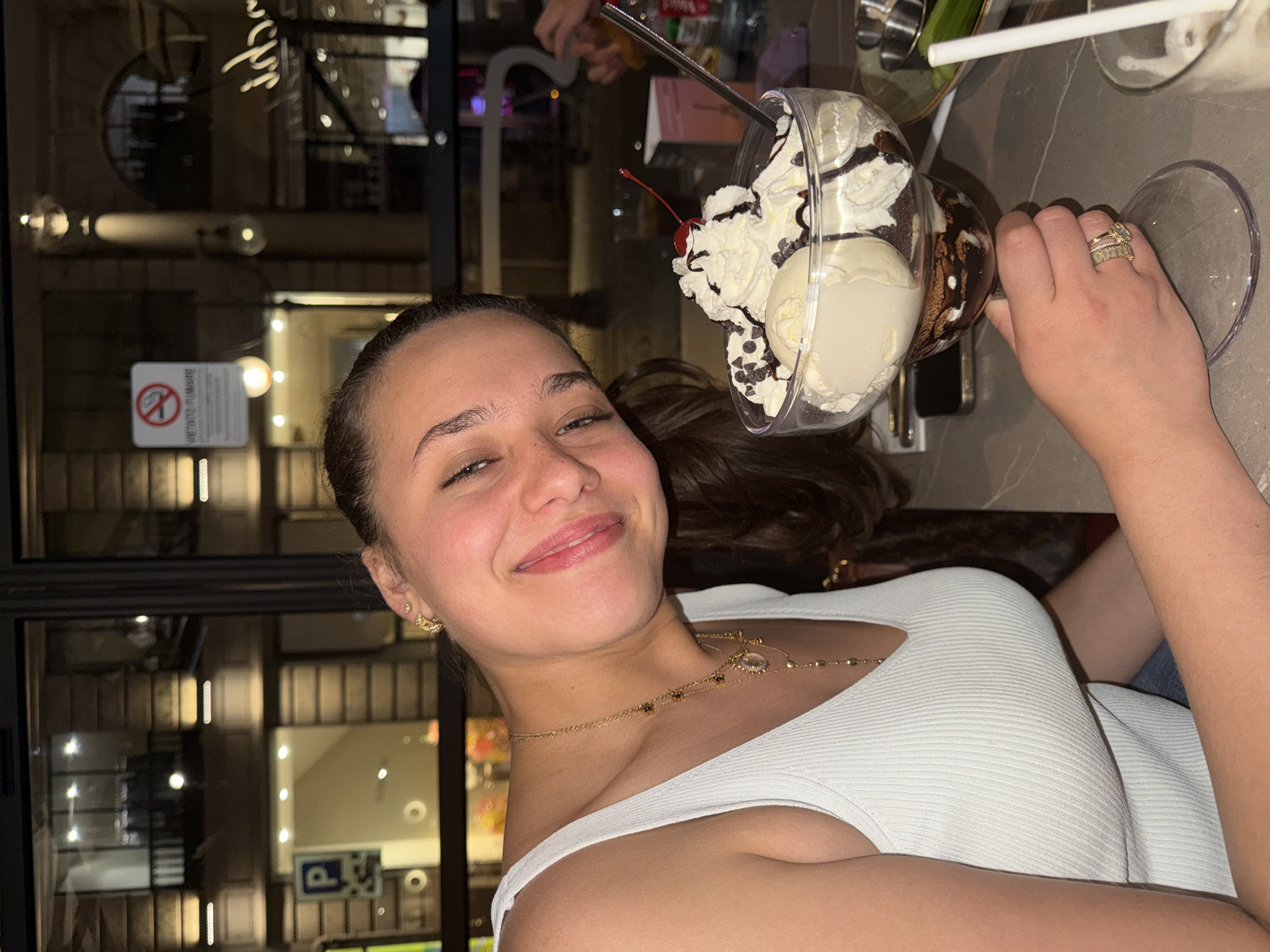 A young woman smiles warmly at the camera, holding a large, ornate sundae glass. She has dark hair tied back, brown eyes, and is wearing a white ribbed tank top, gold stud earrings, and a delicate gold necklace. Her right hand, adorned with several gold rings, grasps the stem of the glass.
The dessert features generous scoops of vanilla ice cream, topped with whipped cream, a drizzle of chocolate syrup, scattered chocolate chips, and a single red maraschino cherry.
The setting is an indoor establishment, likely a cafe or dessert parlor in Milan, Italy, during the evening. The background, seen through a large glass window or partition, is blurred with reflections of indoor and outdoor lights, suggesting an urban environment. A sign prominently displays the universal "No Smoking" symbol and the Italian text "VIETATO FUMARE". Further in the background, a blue square sign with a white 'P' (parking) is partially visible. The woman's broad smile conveys a sense of happiness and enjoyment of her dessert.