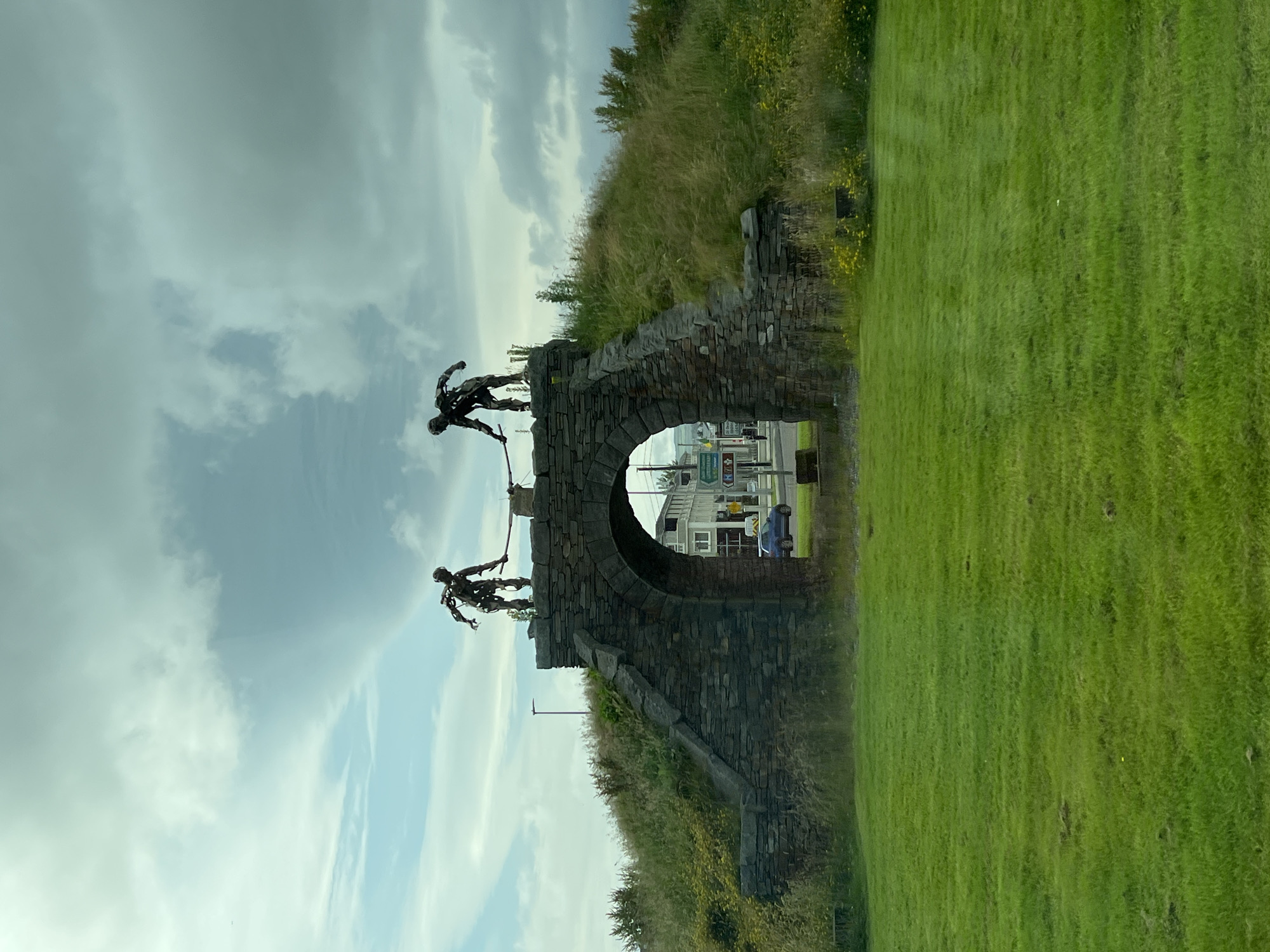 The image depicts an outdoor scene with a stone archway as the central element. Perched on top of the archway are two metal sculptures of figures, seemingly in motion. One figure is leaning forward, appearing to be in mid-stride or leap, while the other is positioned behind it, also in a dynamic pose. The archway itself is constructed from rough-hewn stones and is overgrown with grass and vegetation on its sides, suggesting it's an older structure.
Through the archway, a glimpse of a town or street scene is visible, with buildings, road signs, and a blue car. This provides a sense of location and context, hinting at a connection between the sculptures and a populated area. To the right of the archway, a vibrant green lawn stretches out, meeting a grassy hill topped with wild grasses and some yellow wildflowers. The sky is overcast with light grey and white clouds, suggesting a diffused daylight. The overall mood is one of movement and perhaps resilience, with the figures appearing to be engaged in some form of athletic endeavor or a symbolic journey. The grass suggests a temperate climate, and the presence of vegetation on the arch points towards a season where plant life is abundant. The scene is static, capturing a moment in time, but the sculptures themselves convey a sense of dynamic action.