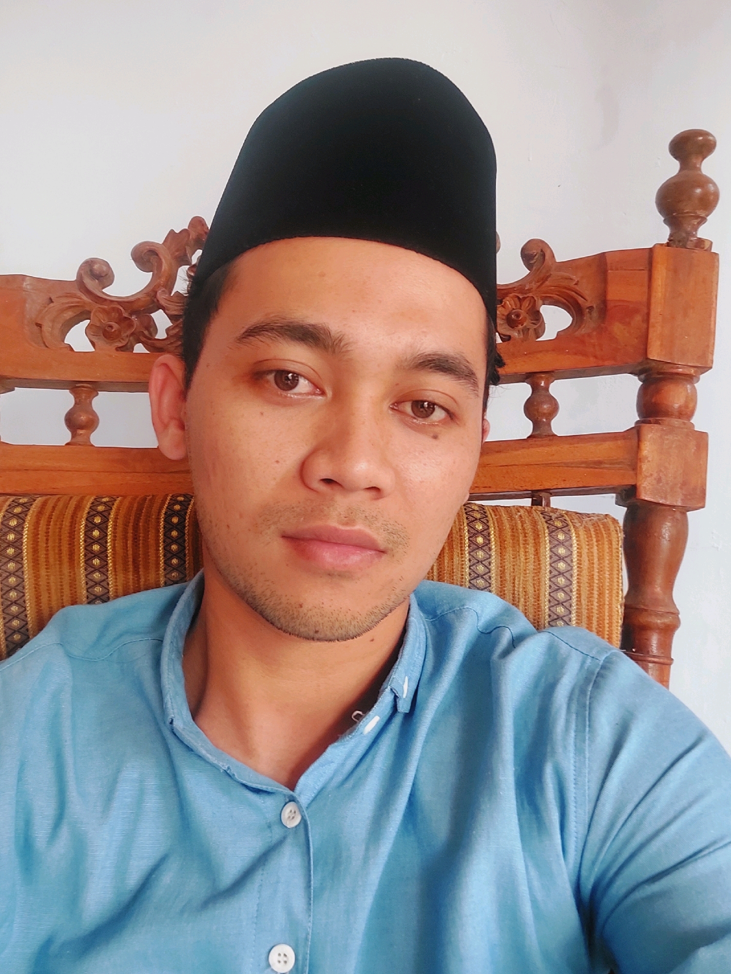A man wearing a black kopiah and a light blue button-up shirt is seated in a wooden chair with an ornately carved backrest and a striped cushion. The man is looking directly at the camera, and his face is centered in the frame. The background is a plain white wall. The scene appears to be indoors.