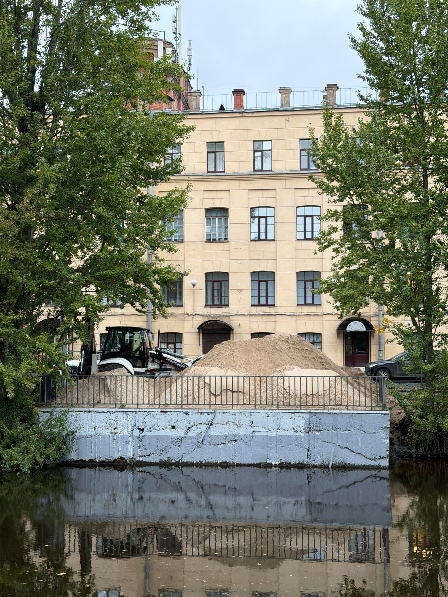 The image shows a construction site next to a canal in Saint Petersburg, Russia. A pile of sand is being excavated by a backhoe, with a fence separating it from the canal. The canal reflects the building behind it, which appears to be an apartment building. The sky is overcast, and there are trees on either side of the frame. The building is light yellow and has several windows. There is a dark-colored car parked on the right side of the image. A person is visible inside the backhoe. The overall scene suggests ongoing construction work in an urban environment.