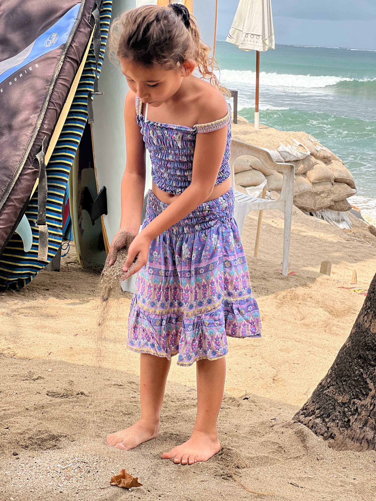 A young girl with curly blonde hair tied back in a ponytail is standing on a sandy beach in Kuta, Indonesia. She is wearing a purple and blue floral print two-piece outfit, consisting of a smocked crop top with off-the-shoulder ruffled sleeves and a matching tiered skirt. Her hands are cupped together, and she is letting sand fall through her fingers. Her bare feet are visible on the sand.
Behind her, a large black and blue surfboard bag stands upright, with the words "SERIES" visible. Several surfboards are leaning against it. In the background, the turquoise ocean with gentle waves is visible, along with a white umbrella and a white plastic chair on a sandbag embankment. The overall scene is a bright, sunny day at the beach.