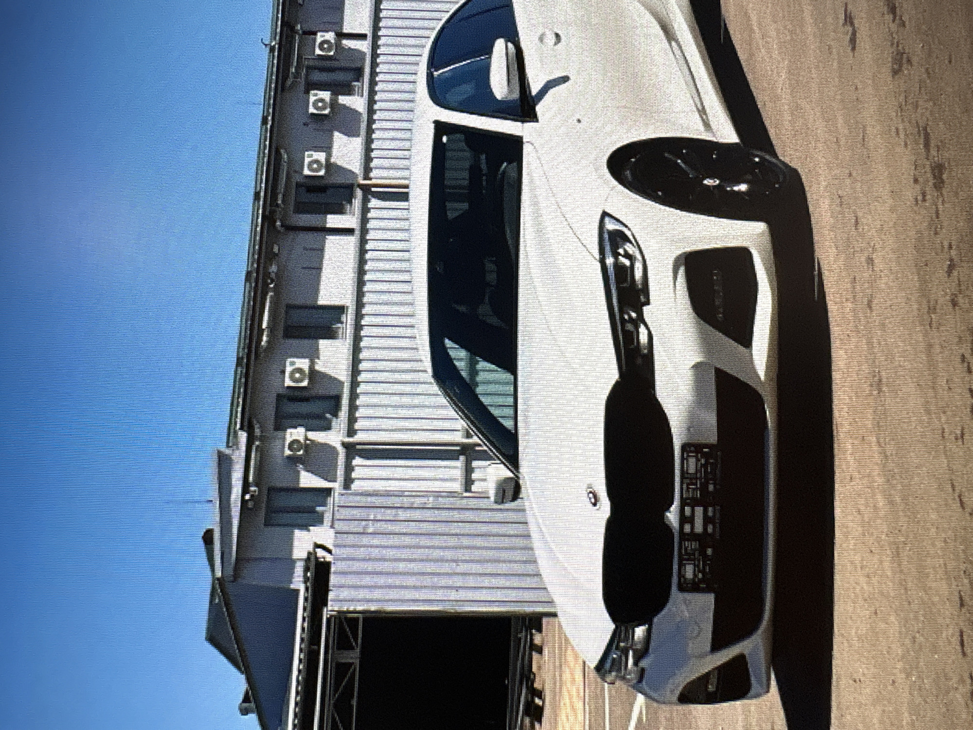 A white BMW sedan is parked in front of a modern industrial building under a clear blue sky. The car's front grille is blacked out, and its headlights are illuminated. The building behind the car features a corrugated metal facade and multiple air conditioning units mounted on the upper wall. The ground in front of the building is paved with asphalt. The scene appears to be a daytime outdoor setting.