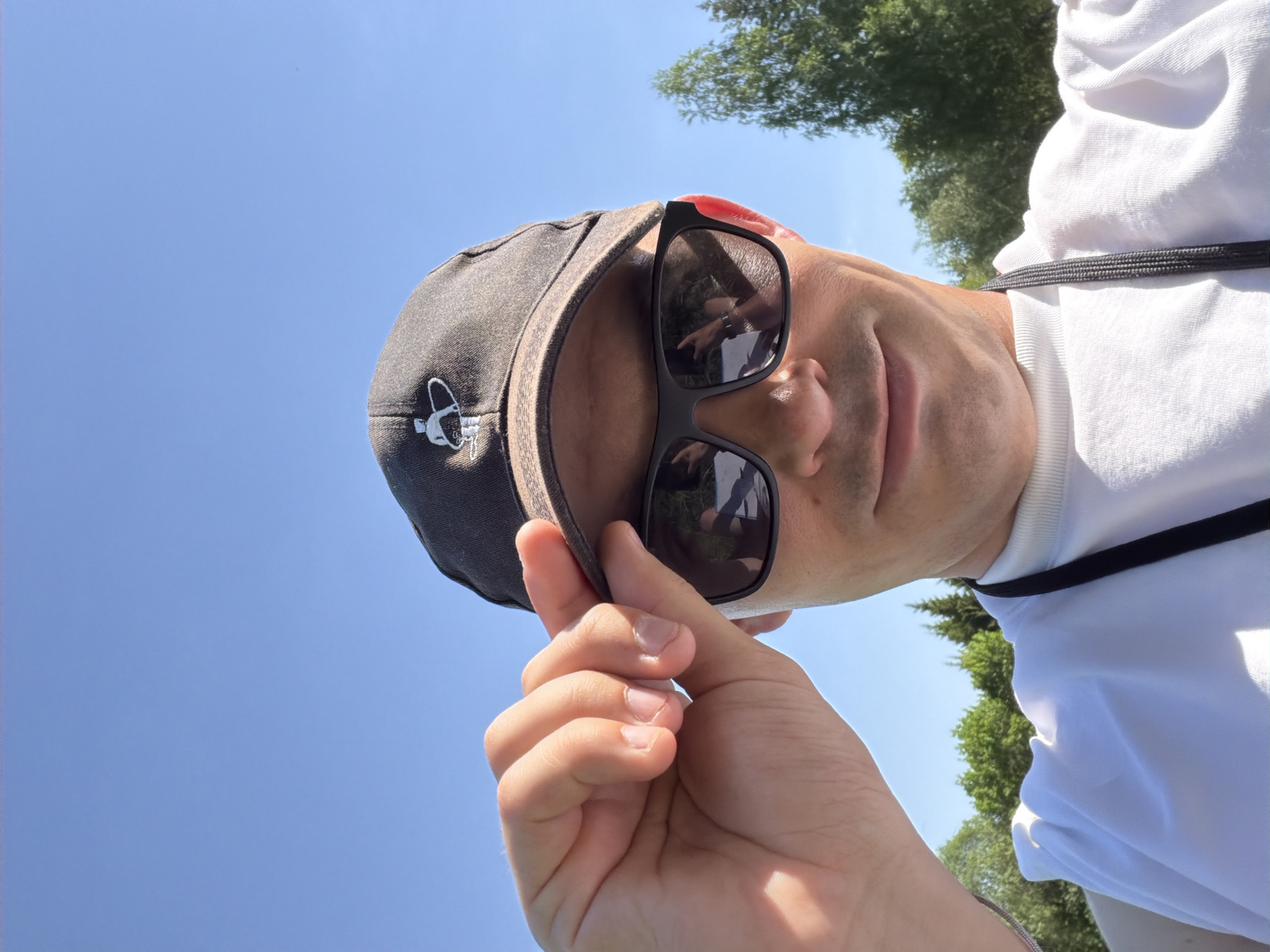 The image depicts a person in Oreshniki, Russia, wearing a white t-shirt, a dark-colored baseball cap with a small white emblem on the front, and sunglasses. The person is holding the brim of the cap with their hand. The background consists of a clear blue sky and some green trees, suggesting an outdoor location. The person appears to be taking a selfie, based on the camera angle and the positioning of their hand. A lanyard hangs around the person's neck.