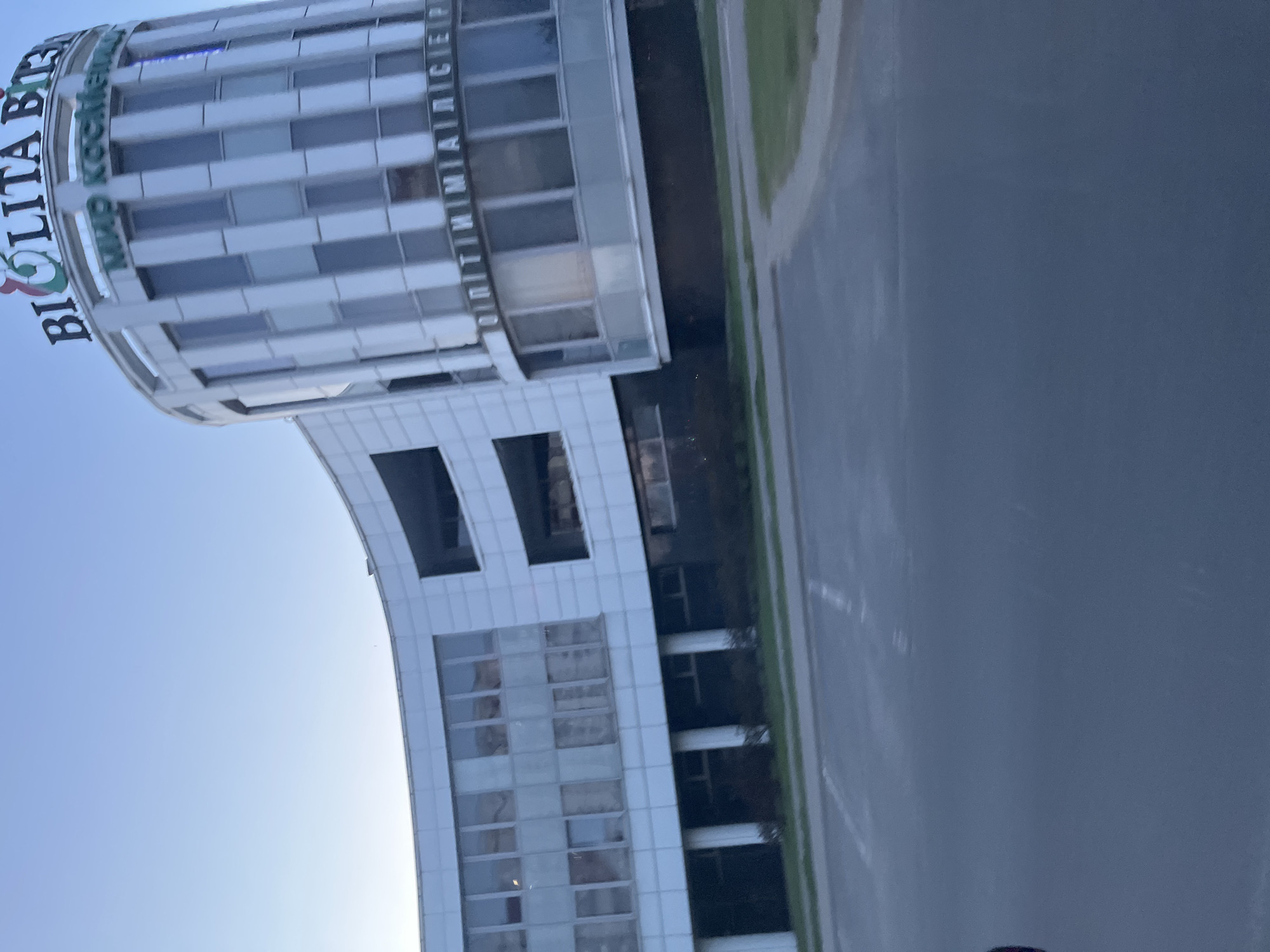The image shows a modern, multi-story building with a curved facade, located in the city of Brest, Belarus. The building features a white exterior with vertical grey panels and large windows. The top of the building is adorned with signage, including what appears to be "BIOLITA BIZ" and "МИР КОСМЕТИКИ" (World of Cosmetics) in Cyrillic. The building is set against a clear blue sky. In the foreground, there is an asphalt surface, possibly a parking lot or road, with some green grass and bushes visible along the edge of the building. The scene appears to be captured during daylight hours.