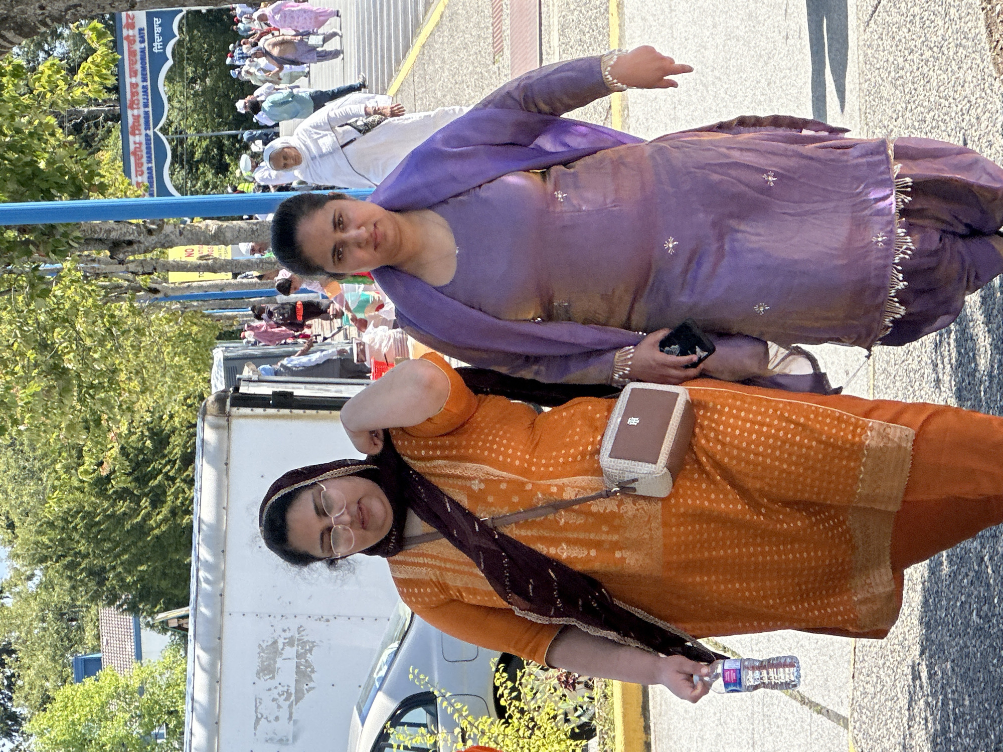 Two women are walking side-by-side outdoors. The woman on the left is wearing an orange patterned salwar kameez and a dark purple dupatta, with glasses and a cross-body purse. She is holding a bottle of water. The woman on the right is wearing a purple salwar kameez and dupatta, with her hands resting on her hips. They are walking on a paved surface with some gravel and are in front of a white trailer. In the background, there are trees, a blue pole, a gate with writing in Punjabi, and a crowd of people, suggesting a public event or gathering. The presence of the "Nidar Hardeep Singh Nijjar Memorial Gate" sign indicates the location is likely the Gurdwara Sahib Dasmesh Darbar in Surrey, British Columbia, Canada.