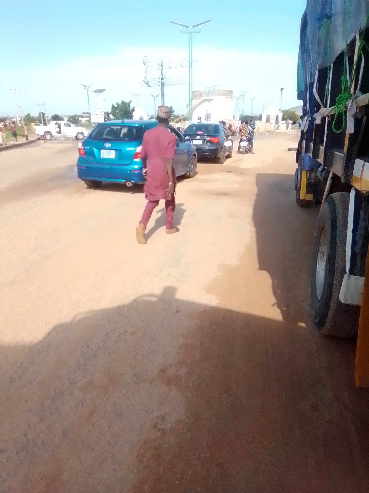 In Kwadon, Nigeria, on a sunny day, a person walks away from the camera on a dirt road. They wear a maroon tunic and pants and a patterned cap. A blue hatchback is parked on the road to their left, and a black sedan is further down on the right. A truck with a green tarp is parked on the far right of the frame. Several other people are visible in the background near a white pickup truck. Solar-powered streetlights are spaced along the road, and in the distance, there are several white buildings with curved roofs, possibly a religious structure.