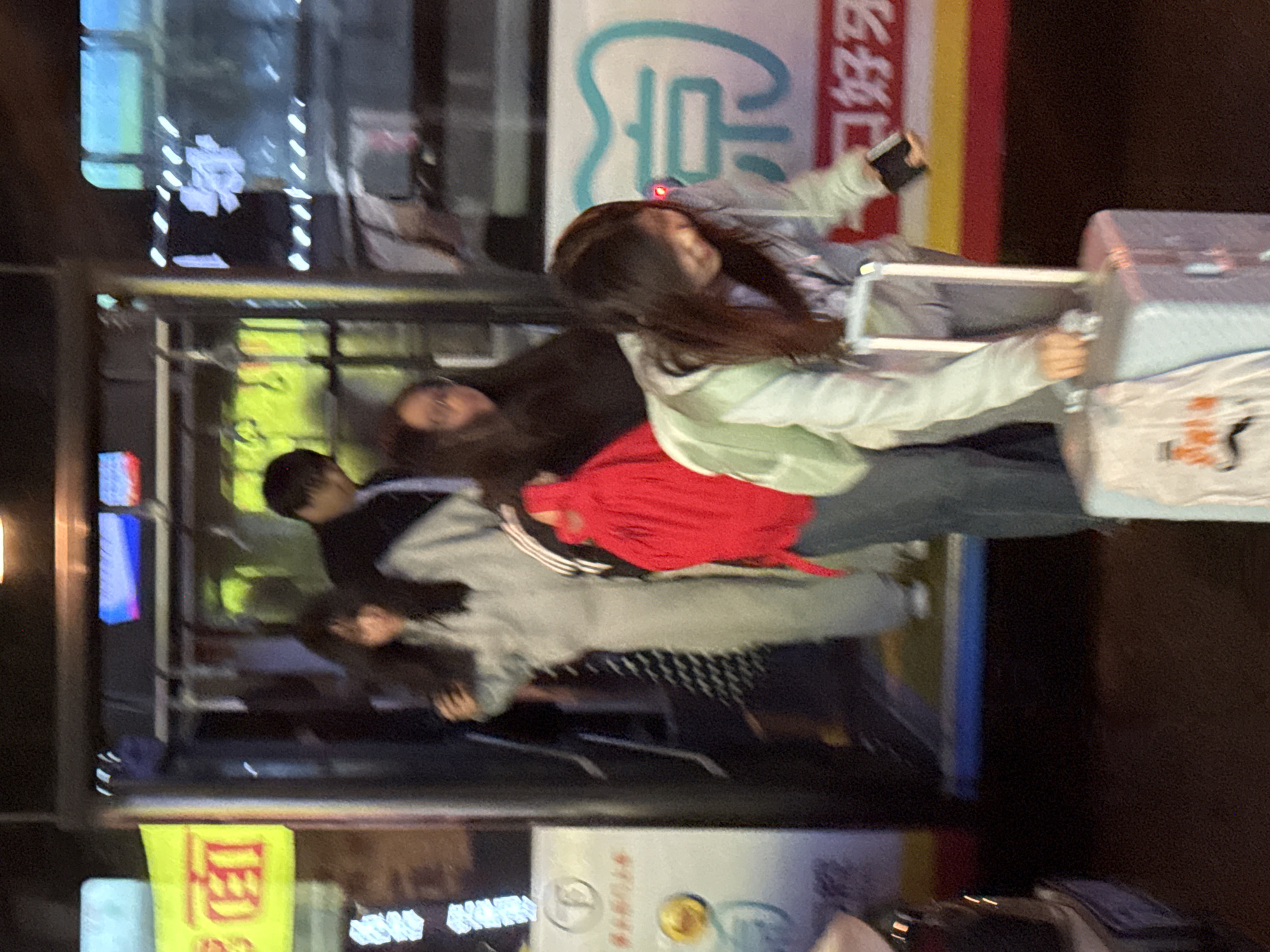 A photo taken in the city of Zhongtun, China, depicts five individuals amidst urban scenery. Three people are standing, one is seated, and another is pulling a suitcase. The person pulling the suitcase is using a phone. The environment shows signs with Chinese characters and various buildings in the background. The photo appears to have been taken at night or in low lighting conditions.