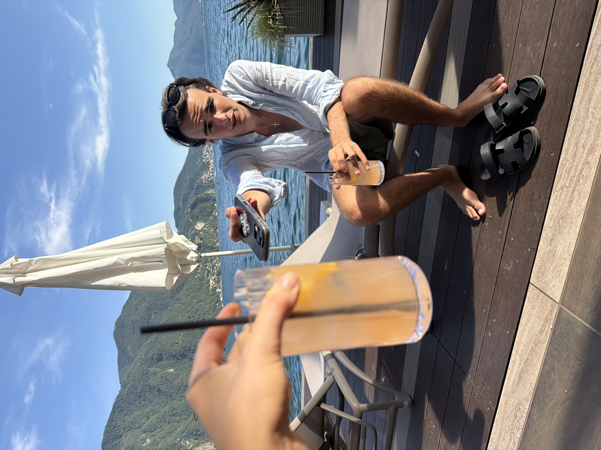 A young man with dark hair and sunglasses perched on his head sits on a lounge chair outdoors, holding a phone and a drink. He wears a light blue, unbuttoned shirt and dark shorts. His bare feet are visible, with black sandals on the wooden deck beside him. In the foreground, a hand holds a tall glass of an orange-colored beverage with a straw. Behind the man, a body of water, mountains, and a white umbrella are visible, suggesting a location in Lugano, Switzerland, likely at a lakeside establishment. The sky is blue with scattered clouds. The overall scene depicts a relaxed outdoor setting on a sunny day.