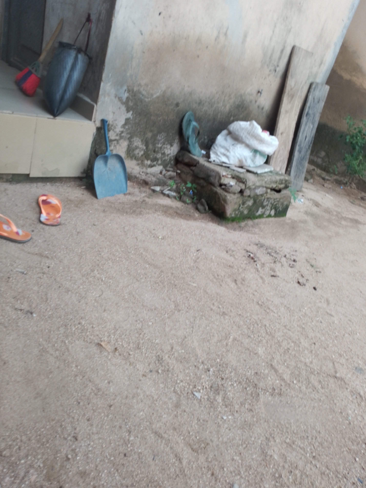 In Jauro Yinu, Nigeria, a sandy outdoor area features several objects. To the left, a red and black broom rests against a cream-colored structure. Next to the broom is a large, dark gray bag. A blue shovel stands upright in the sand near the wall. In the middle ground, a white bag or bundle is placed on a stone ledge. Beside the bundle are two pieces of weathered wood leaning against the wall. On the sandy ground to the left, two orange flip-flops are visible. A single green flip-flop lies on the stone ledge near the white bundle. The wall in the background appears aged and worn.